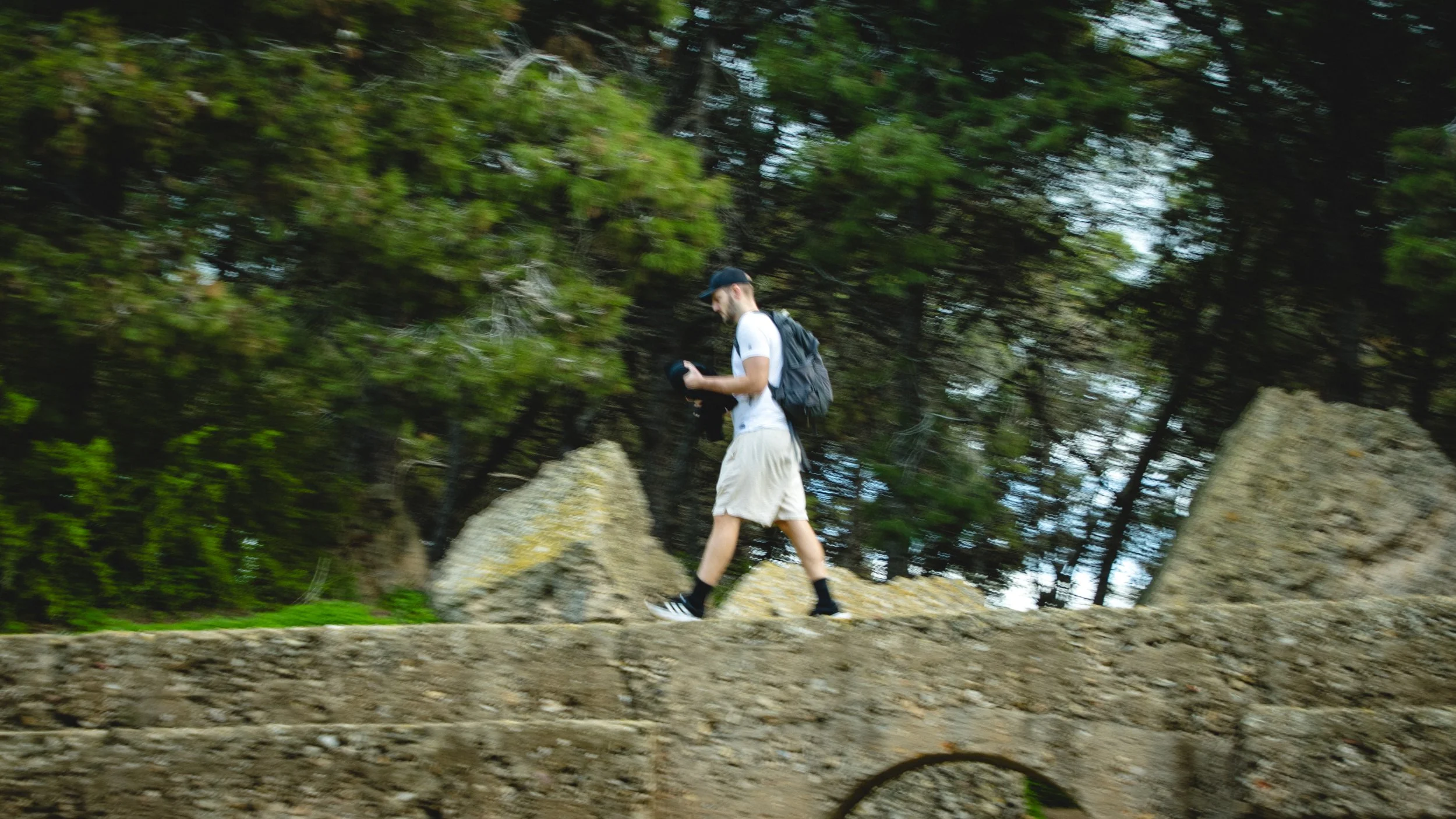 A man wearing a white t-shirt, beige shorts, black socks, and sneakers walks on a rocky trail in a forested area. He is carrying a backpack and looking at a device in his hands.
