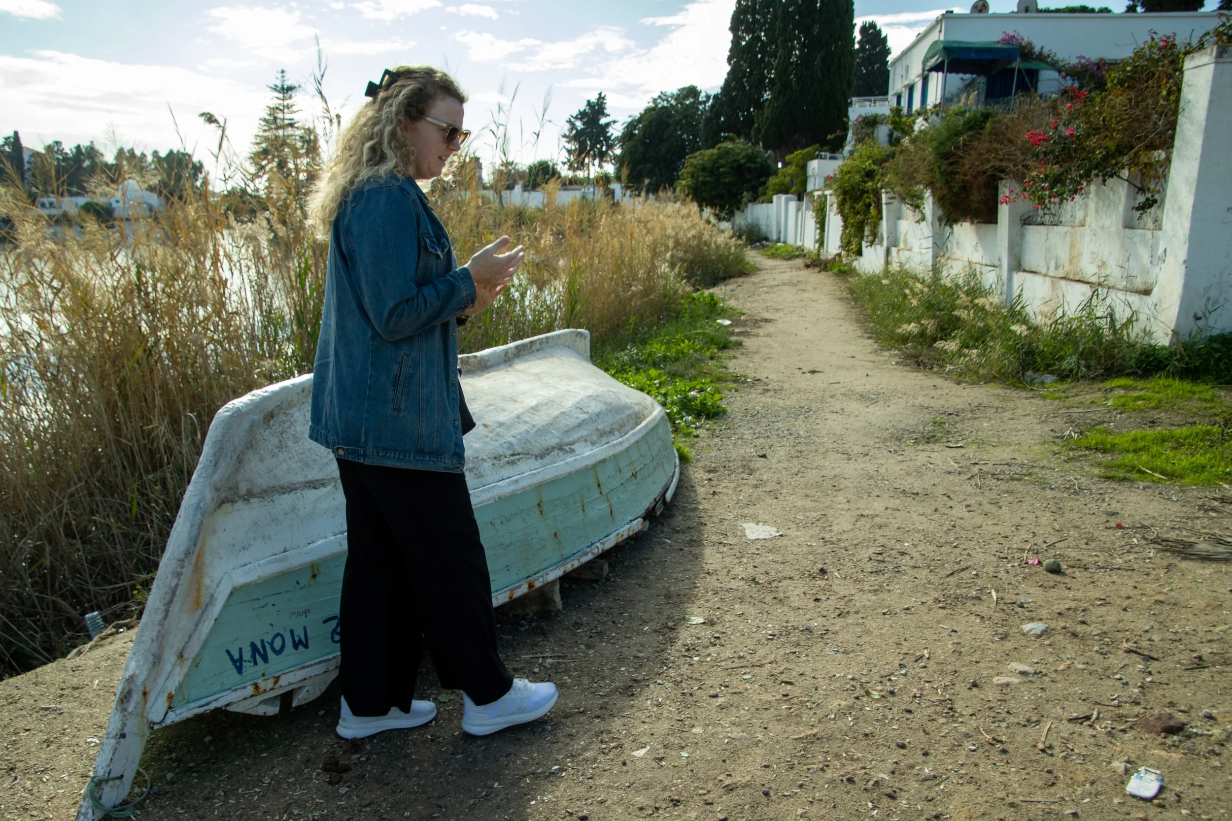 A woman with curly blonde hair, sunglasses, a denim jacket, black pants, and white sneakers is standing beside an upside-down boat on a dirt path. She is looking at her phone. The setting appears to be an outdoor area near residential buildings with 