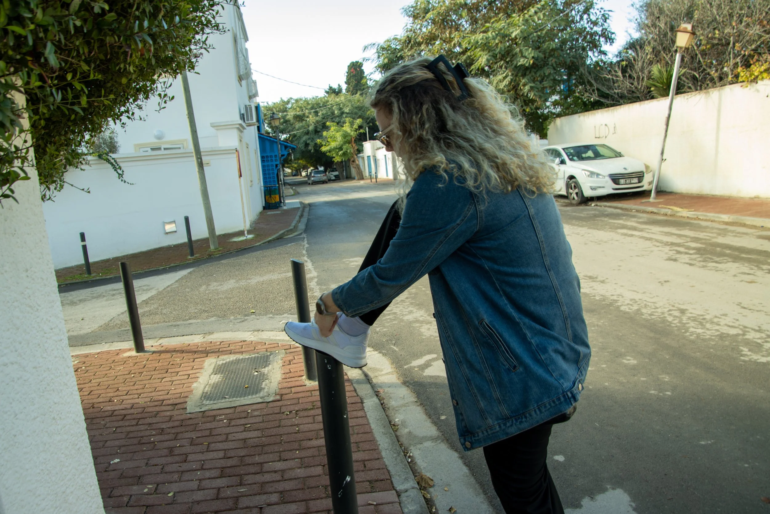 A woman with curly blond hair wearing glasses, a denim jacket, and black pants is stretching her leg on a bicycle rack along a city street.
