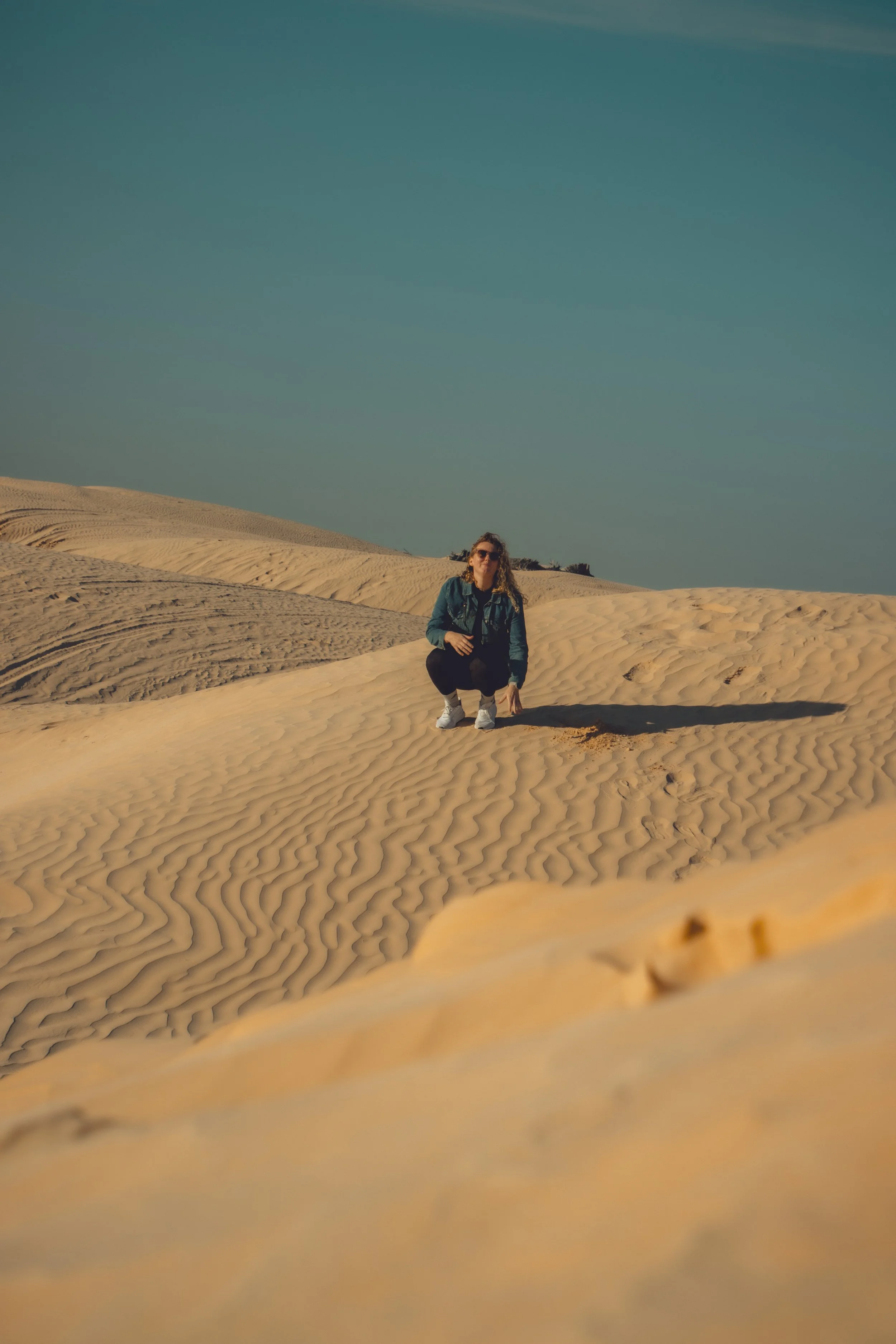 A woman with sunglasses and a denim jacket crouching in sand dunes under a clear blue sky.