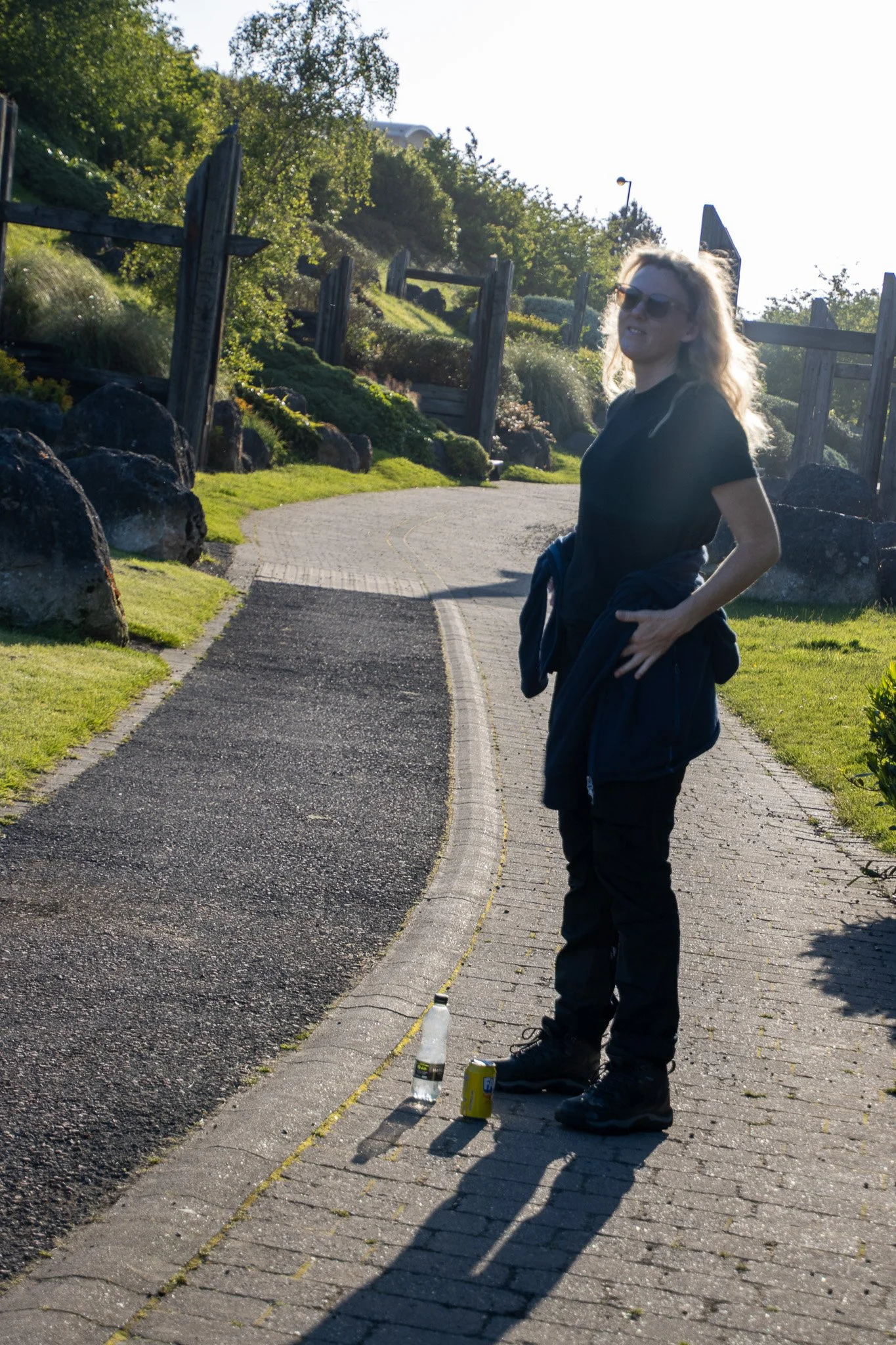 A woman with long blonde hair, sunglasses, and black clothing standing on a paved path outdoors during daylight, with drinks and snacks on the ground in front of her, and greenery and rocks in the background.