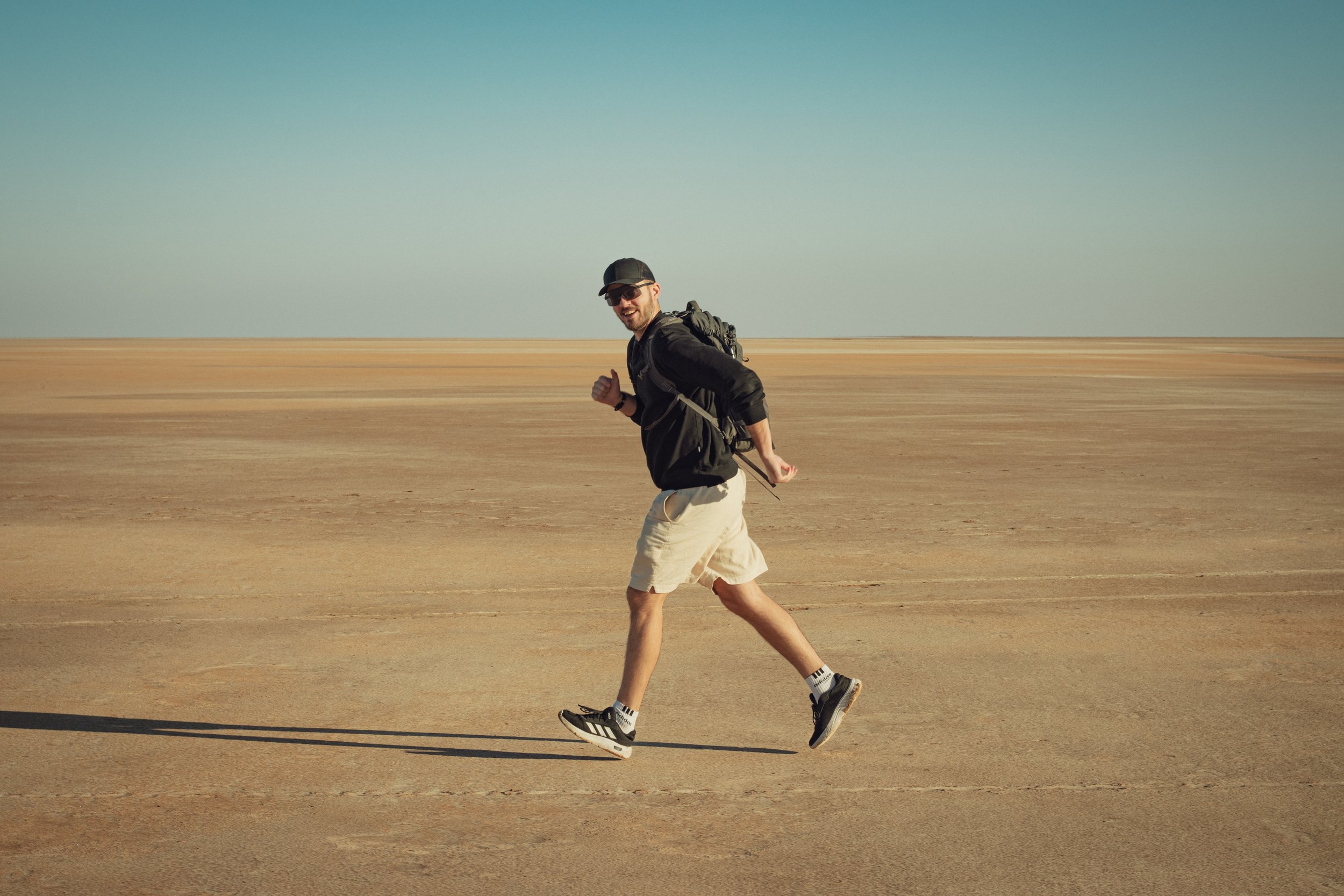 A man running across a vast, flat, desert-like landscape with a clear blue sky above. He is wearing sunglasses, a black cap, a black jacket, khaki shorts, and athletic shoes, and is smiling at the camera.