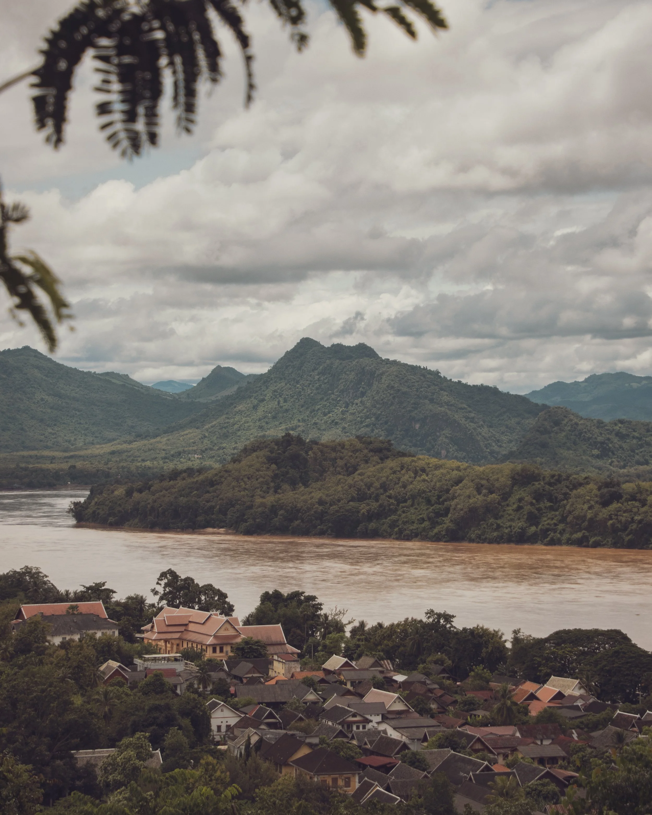 A scenic view of a hilly landscape with dense green vegetation, a river flowing through the valley, and a residential area with houses featuring red and gray roofs in the foreground. The sky above is cloudy with a thick layer of gray clouds.