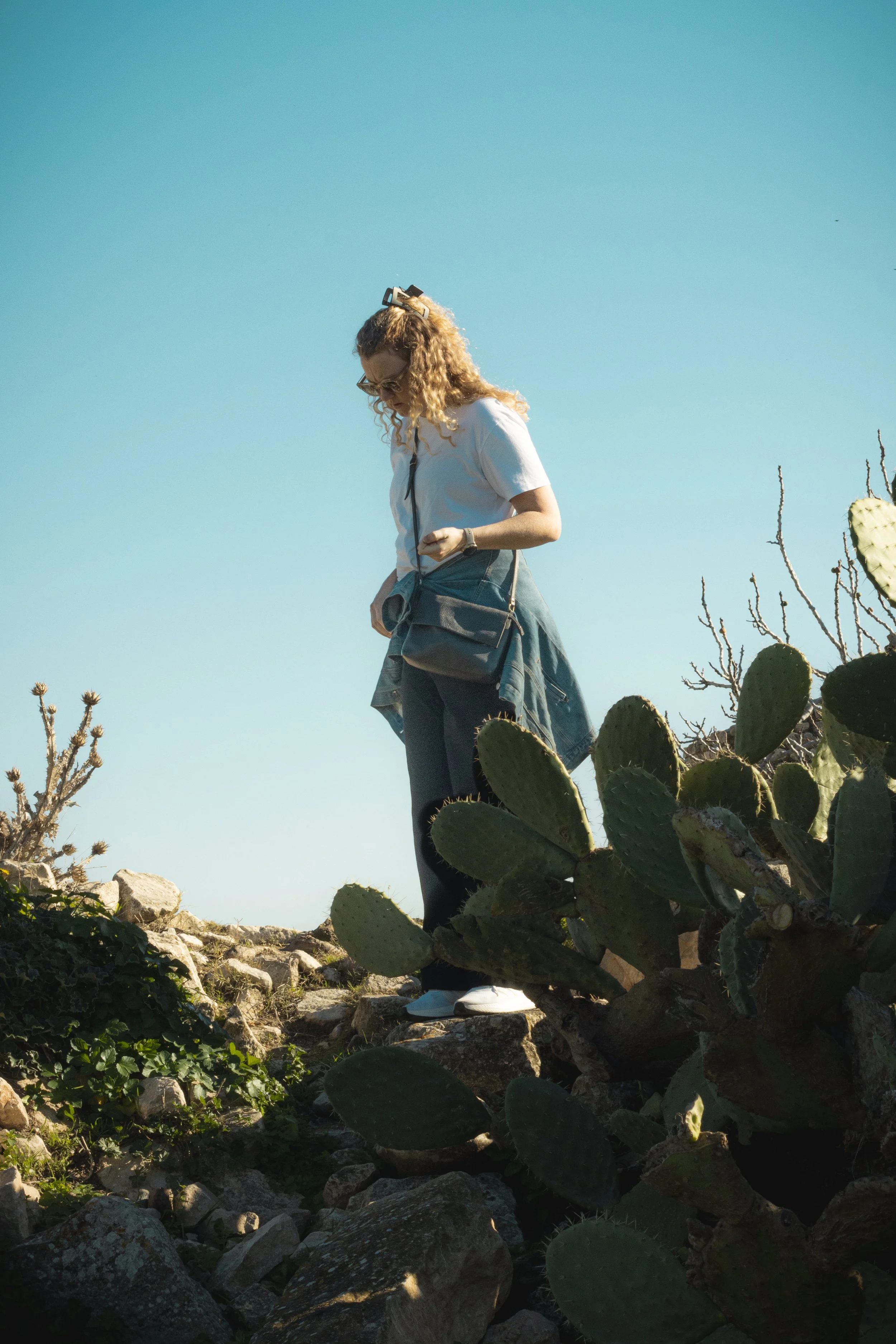 A woman with curly blonde hair and glasses walking amidst desert plants and cacti on rocky terrain under a clear blue sky.