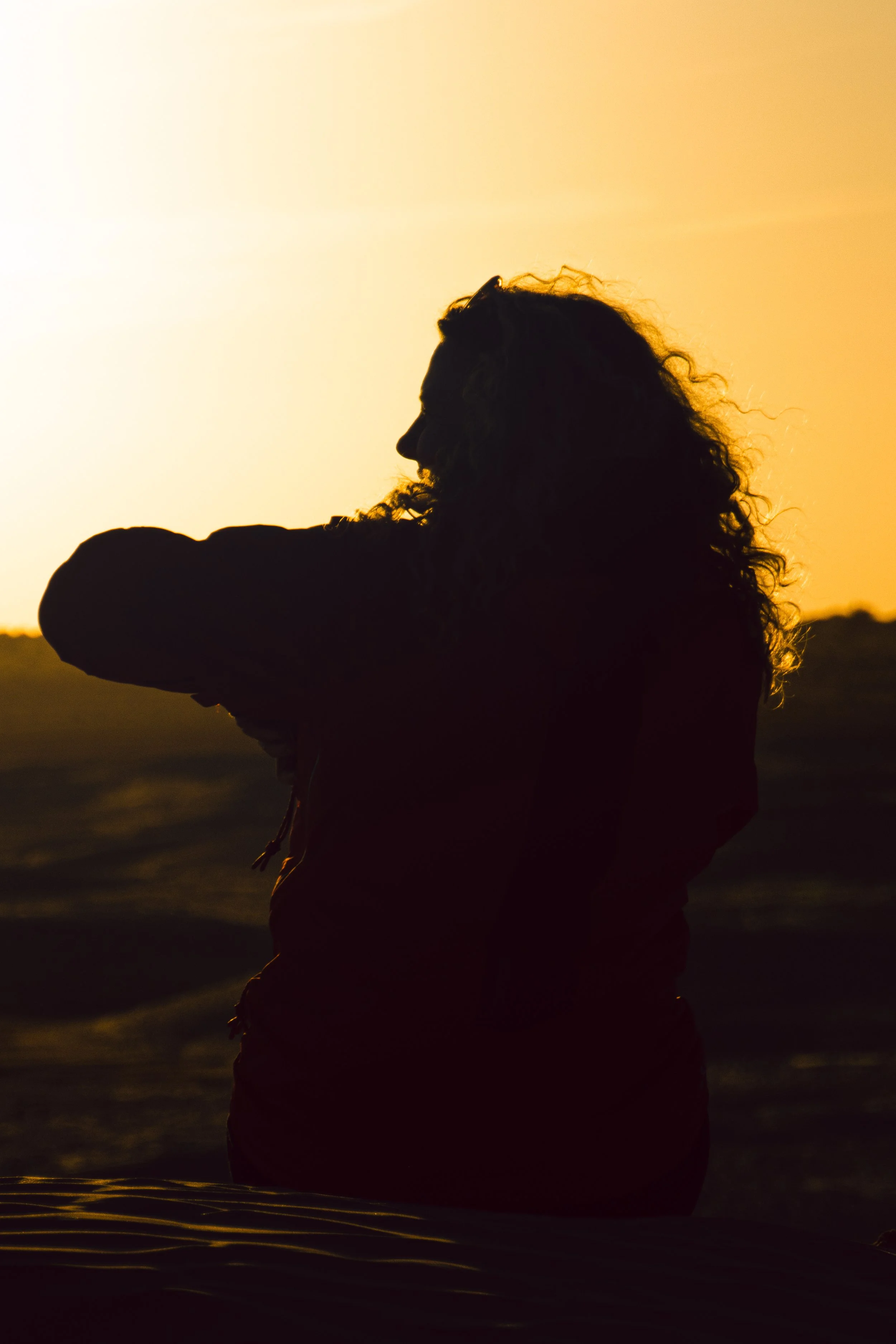 Silhouette of a woman with curly hair at sunset or sunrise near water, with a warm orange sky.