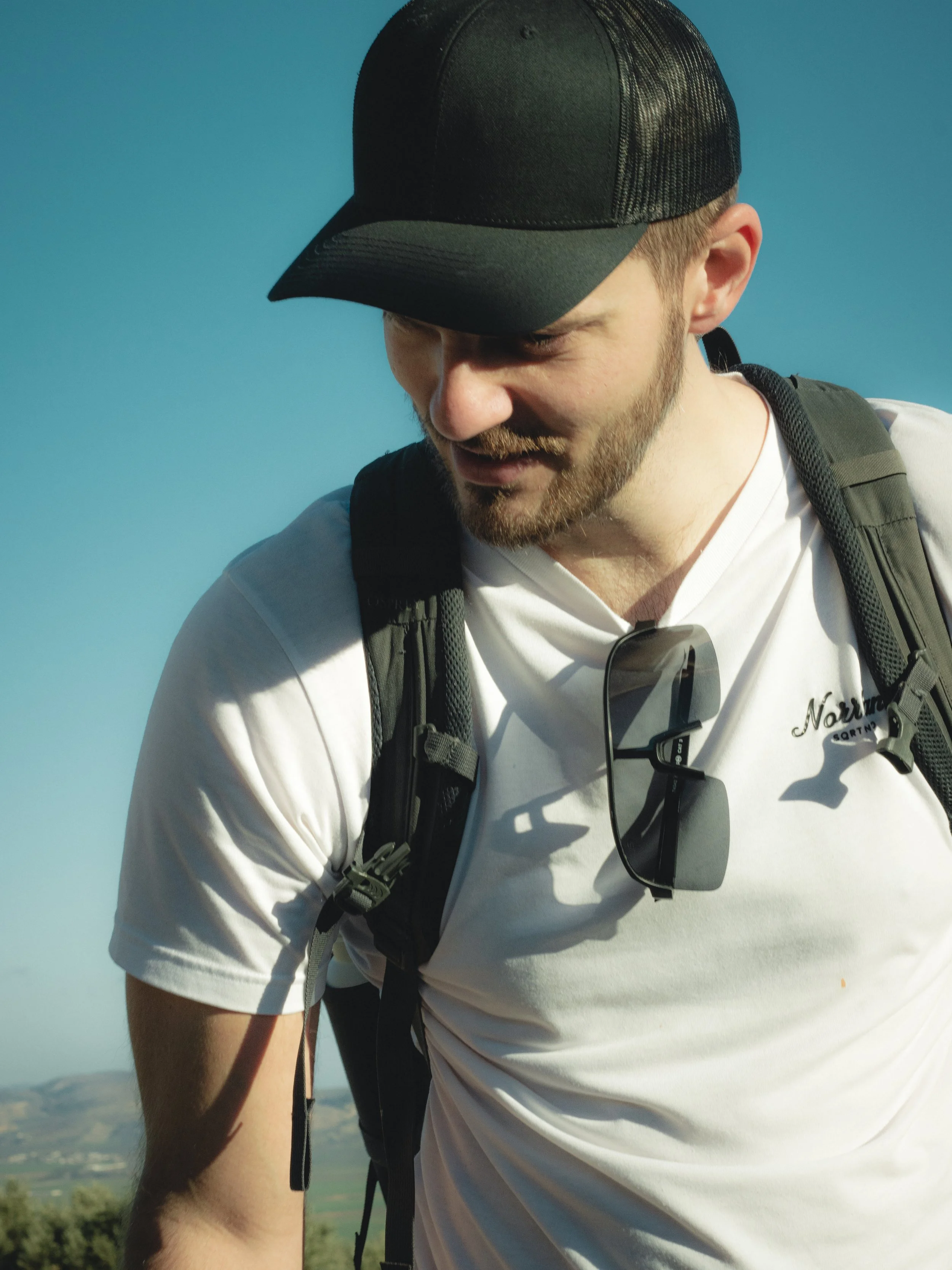 A man wearing a black baseball cap, white T-shirt with a logo, black sunglasses hanging from his shirt collar, and a backpack, looking downward with a background of blue sky and distant green landscape.