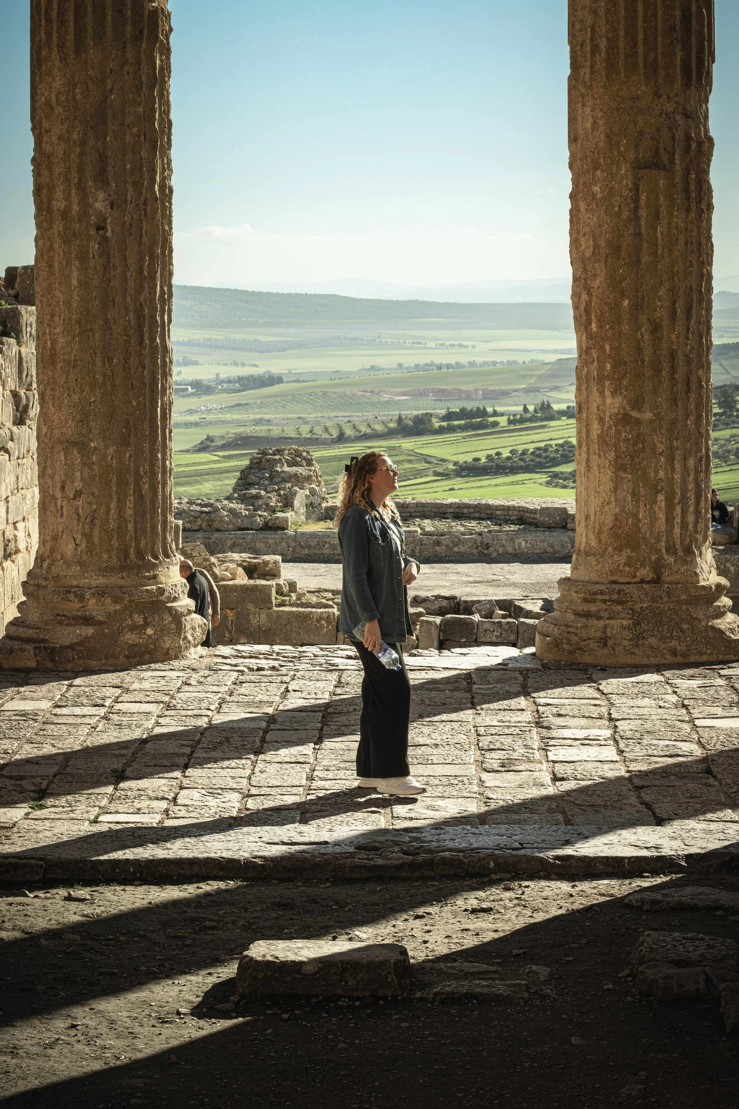 Woman in denim jacket and black pants standing in ancient stone structure with large columns, overlooking lush green landscape and distant hills