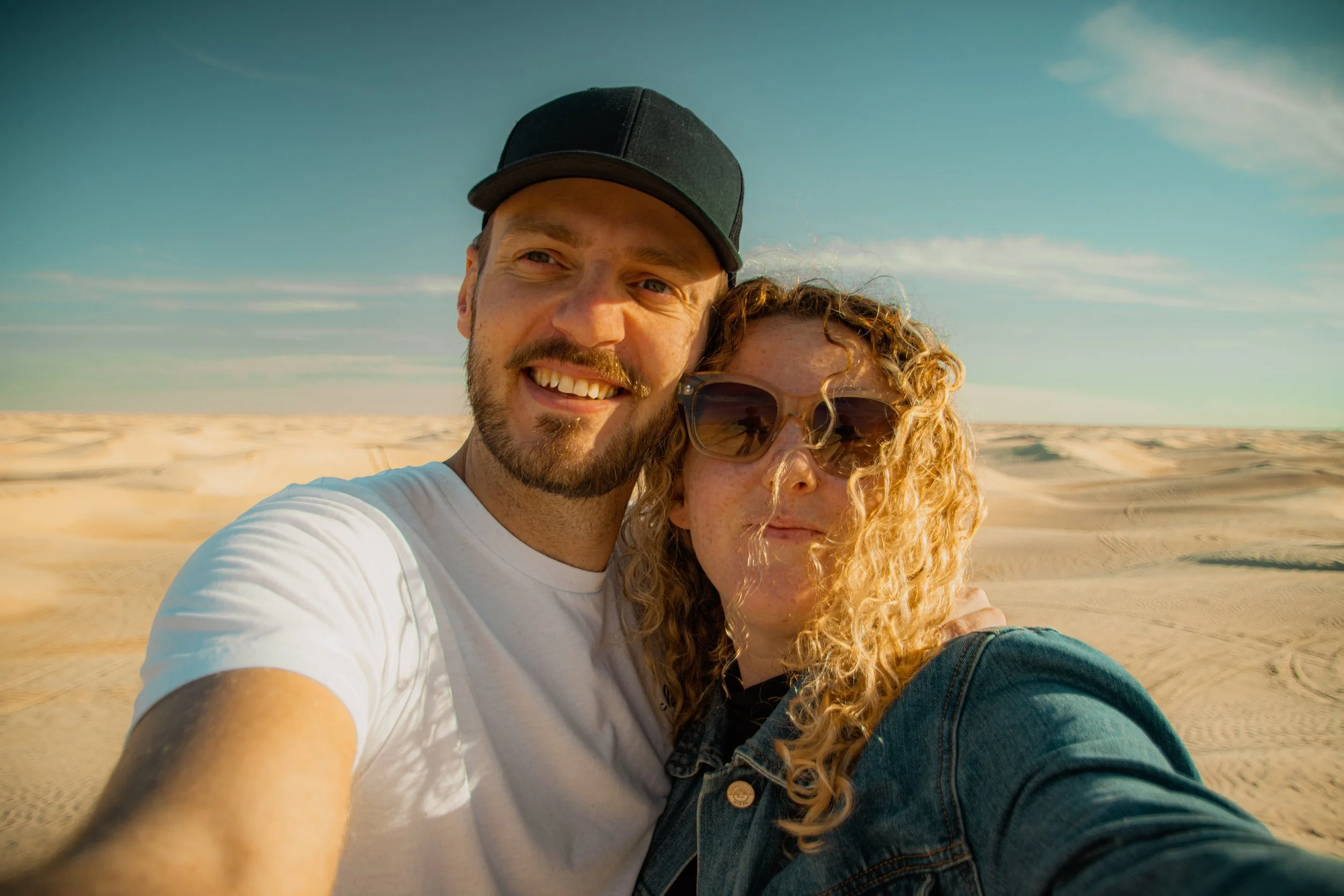 A close-up selfie of a smiling man with a beard wearing a black cap and a woman with curly hair wearing sunglasses, standing on a sandy beach with a clear blue sky in the background.