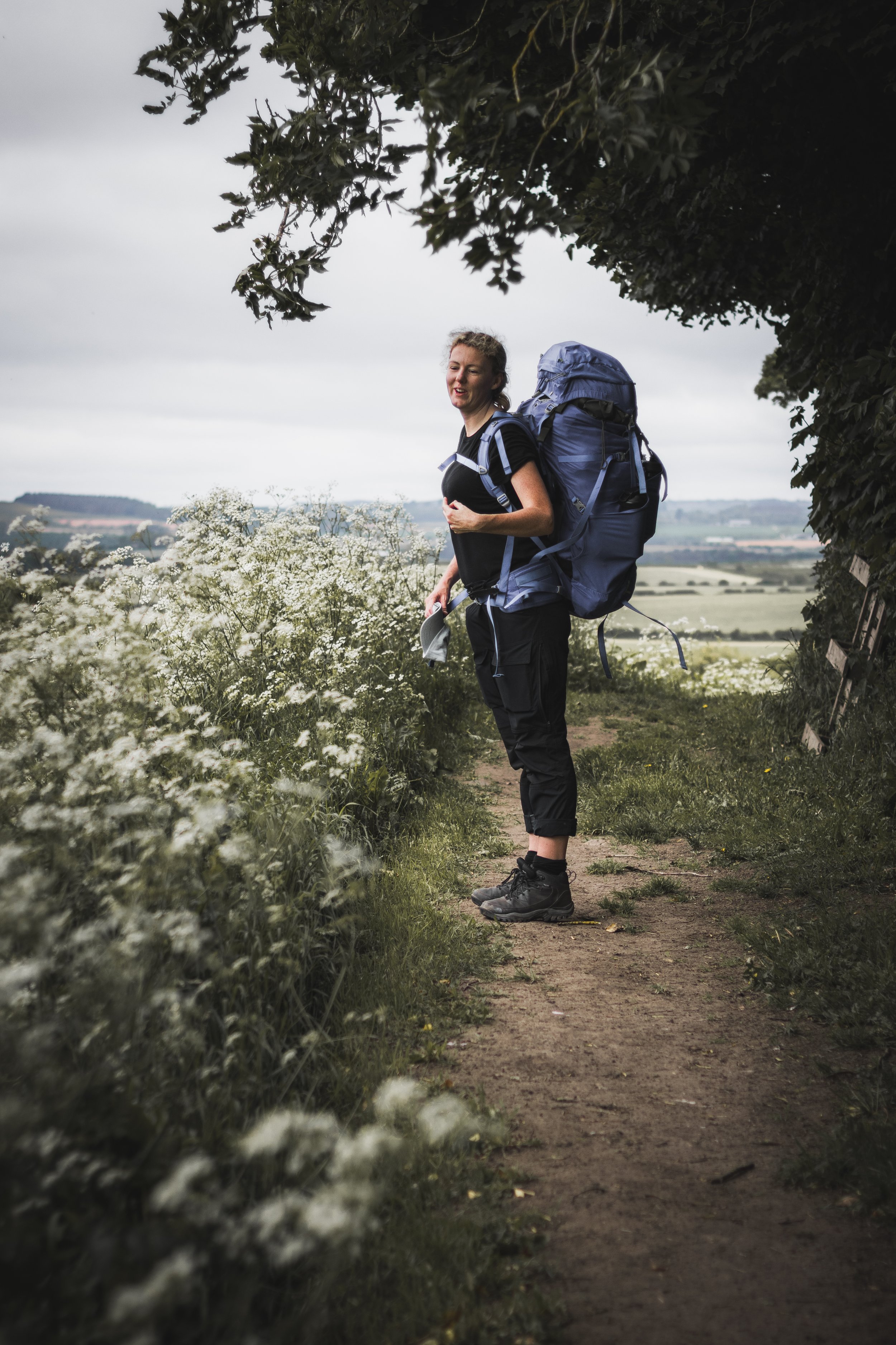 A woman with a large blue backpack standing on a narrow dirt path next to white wildflowers, with rolling hills and cloudy sky in the background.