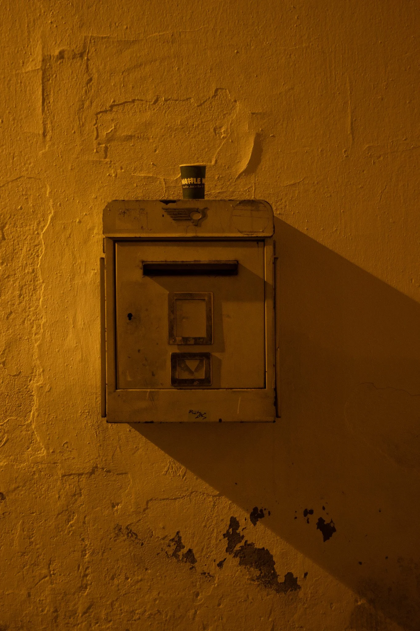 A beige, vintage mailbox mounted on a yellow, textured wall with peeling paint. A black coffee cup labeled "WAFFLE" is placed on top of the mailbox. The lighting has a warm, orange hue.
