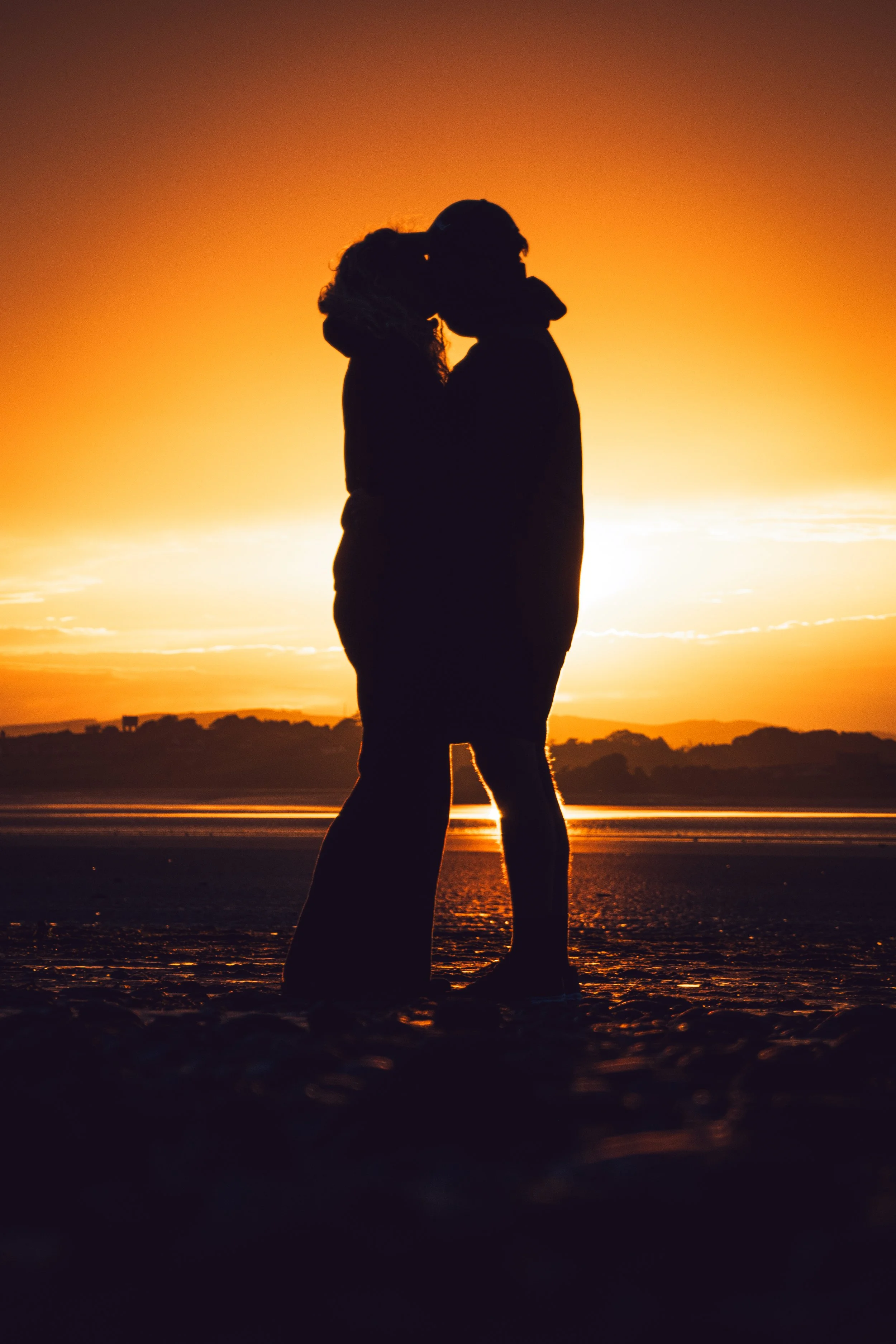 Silhouettes of a couple kissing by the water at sunset or sunrise with an orange and yellow sky.