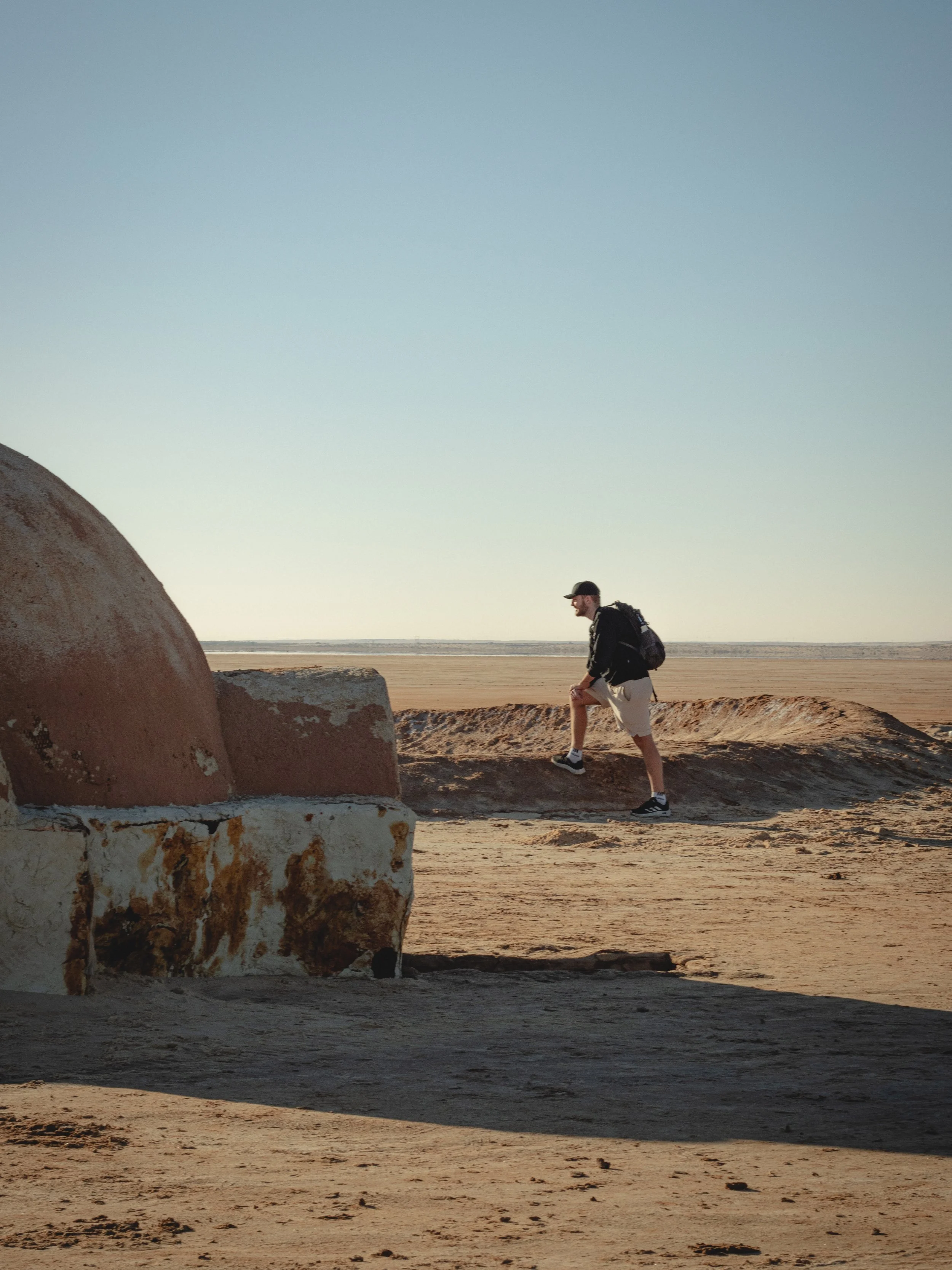A man with a backpack walking in a desert landscape with large rocks, a flat expanse, and a clear sky at sunset