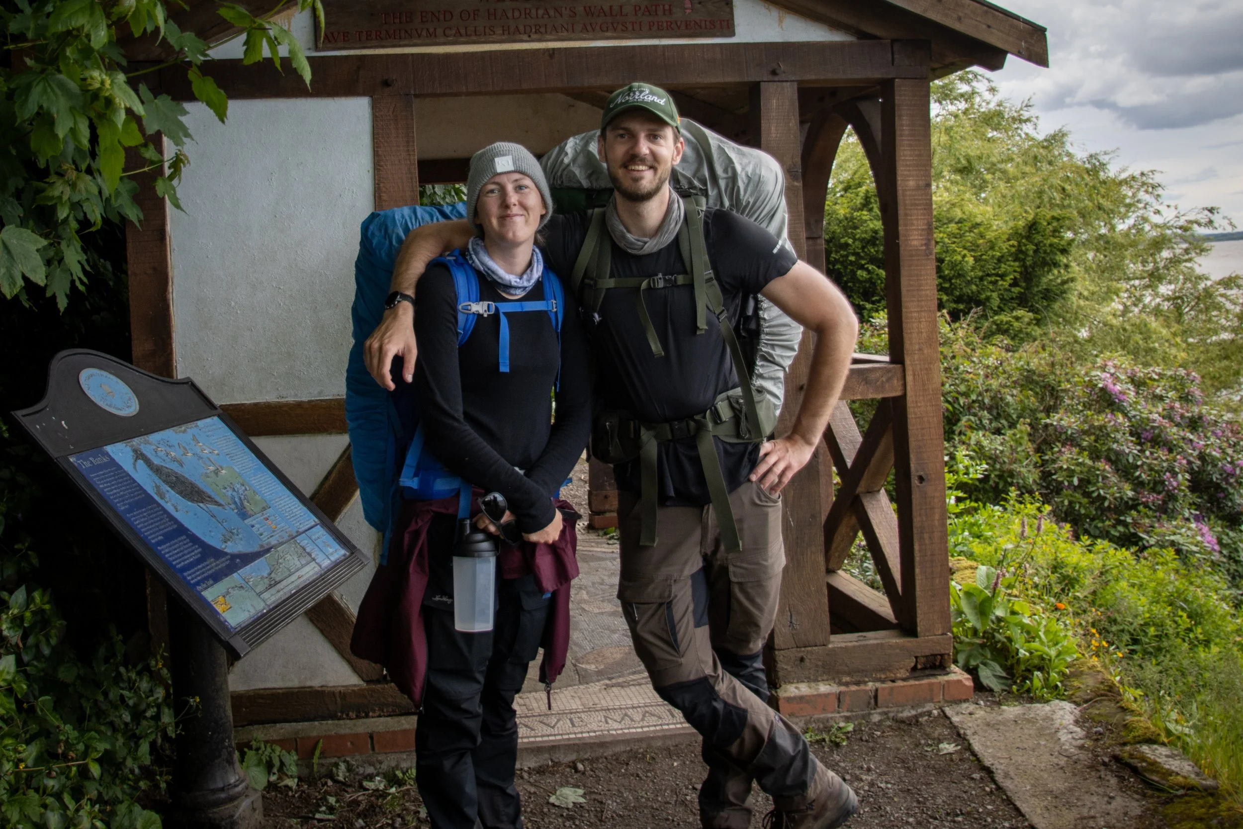 A smiling man and woman with backpacks standing together outdoors near a wooden structure with a sign, surrounded by greenery and flowers.