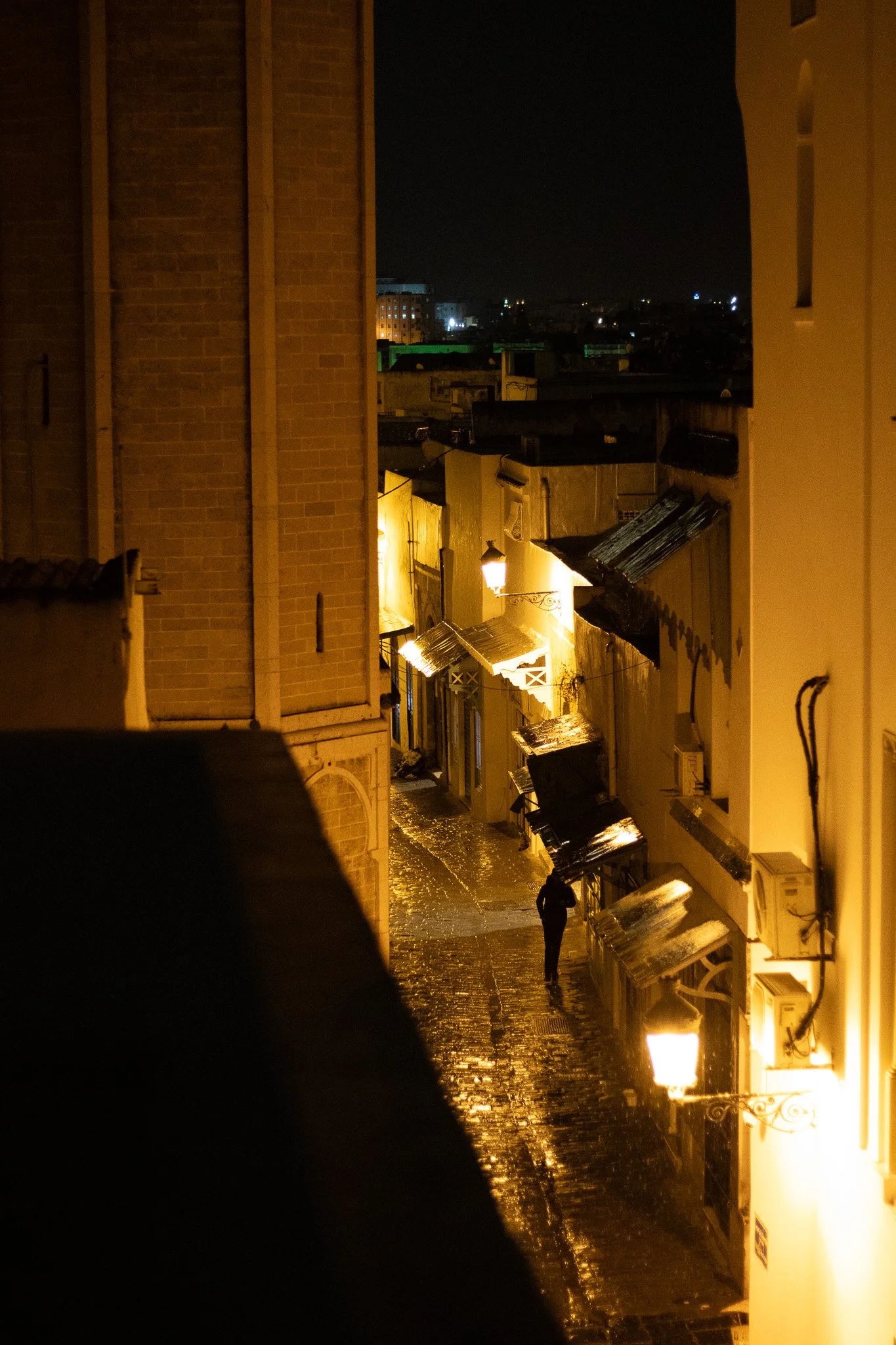 Nighttime street scene with cobblestone road, illuminated by warm yellow lamps, featuring a person walking and buildings on both sides, some with awnings, and a view of city lights in the background.