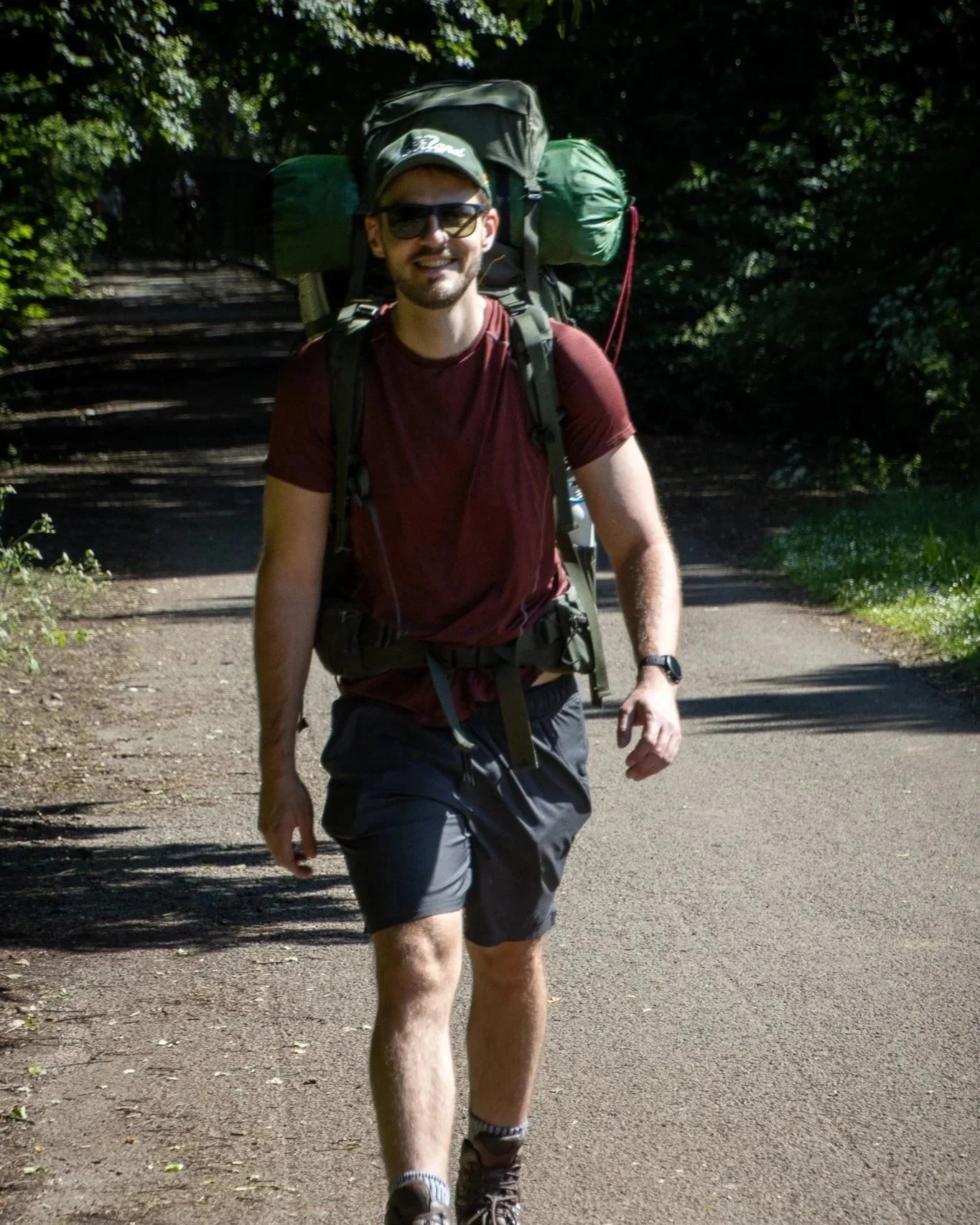 A man hiking on a forest trail, carrying a large backpack and wearing sunglasses, a cap, and outdoor clothing.