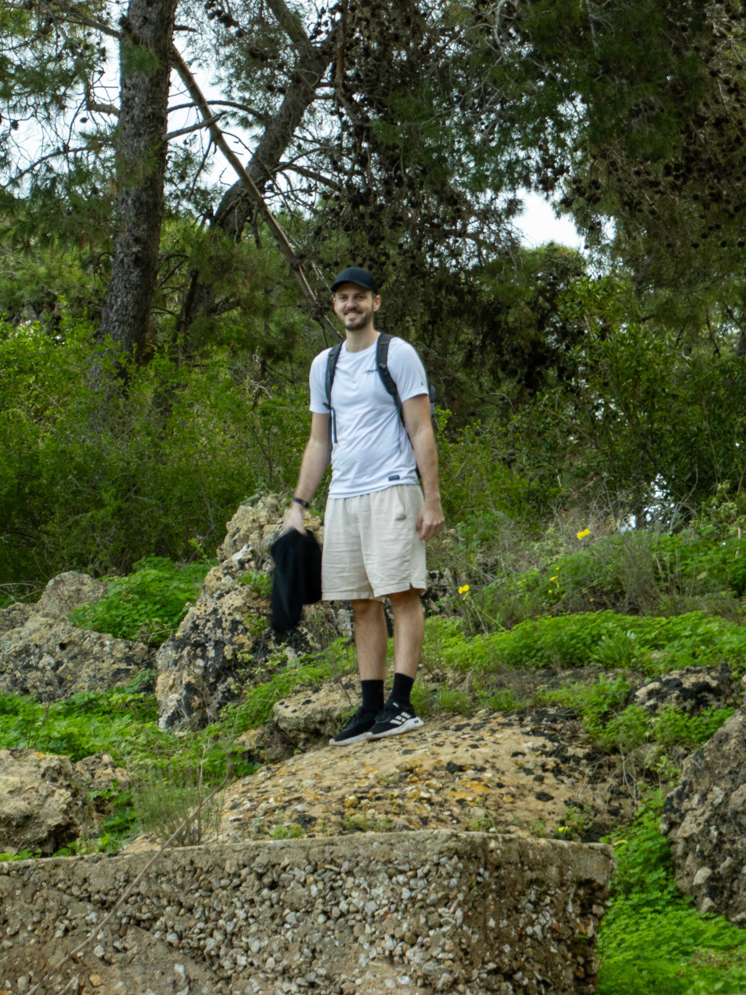 A young man in a white t-shirt, beige shorts, black cap, and sneakers standing on uneven rocky terrain in a forested area, smiling at the camera.