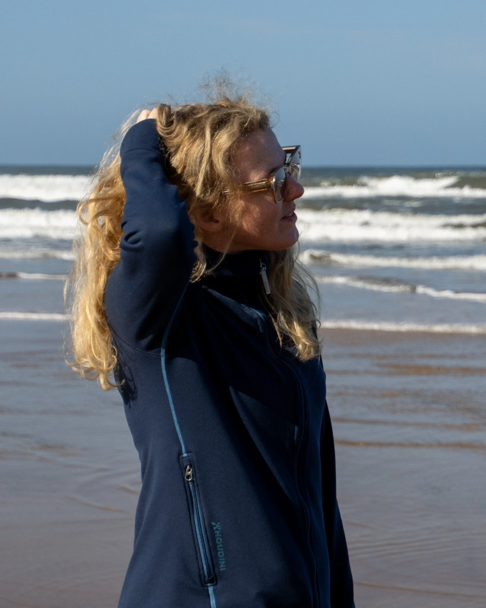 A woman with long wavy blond hair wearing glasses and a dark jacket standing outdoors near the ocean, with waves in the background.