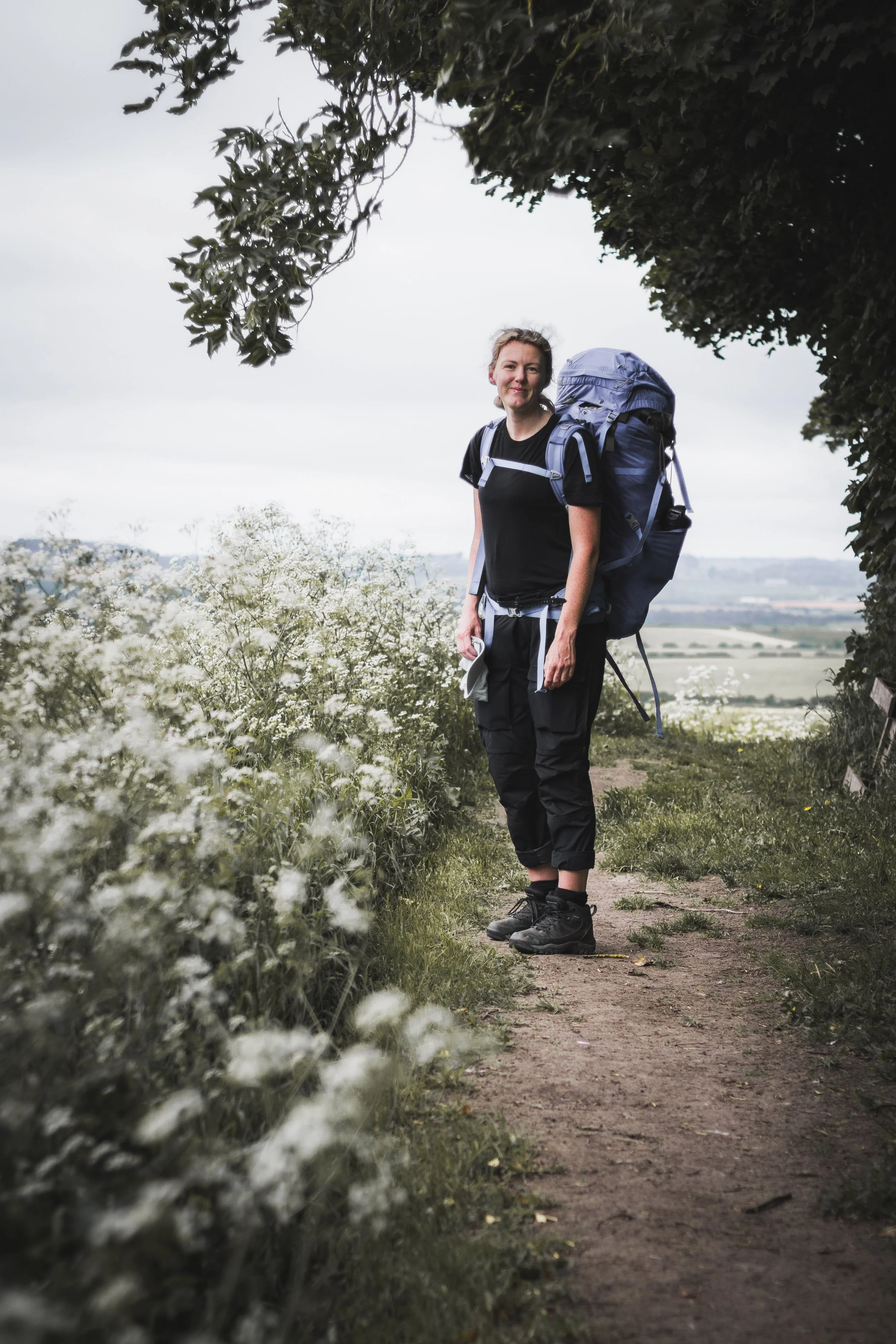 A woman with a backpack standing on a trail in a countryside setting, surrounded by tall flowering plants and trees, with fields and a cloudy sky in the background.