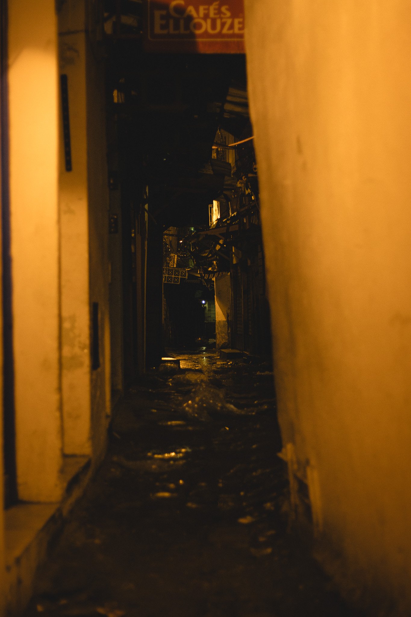 Dark alleyway with wet pavement between yellow walls, dimly lit at night, with a sign reading 'CAFES ELLOUZE' in the background.