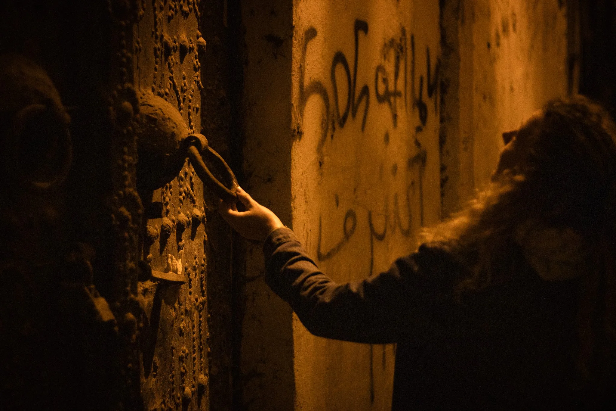 A person with long hair, wearing a jacket, is reaching out to touch a large rusty lock on a weathered metal door or gate that has graffiti on it. The scene is dimly lit with warm, orange lighting.