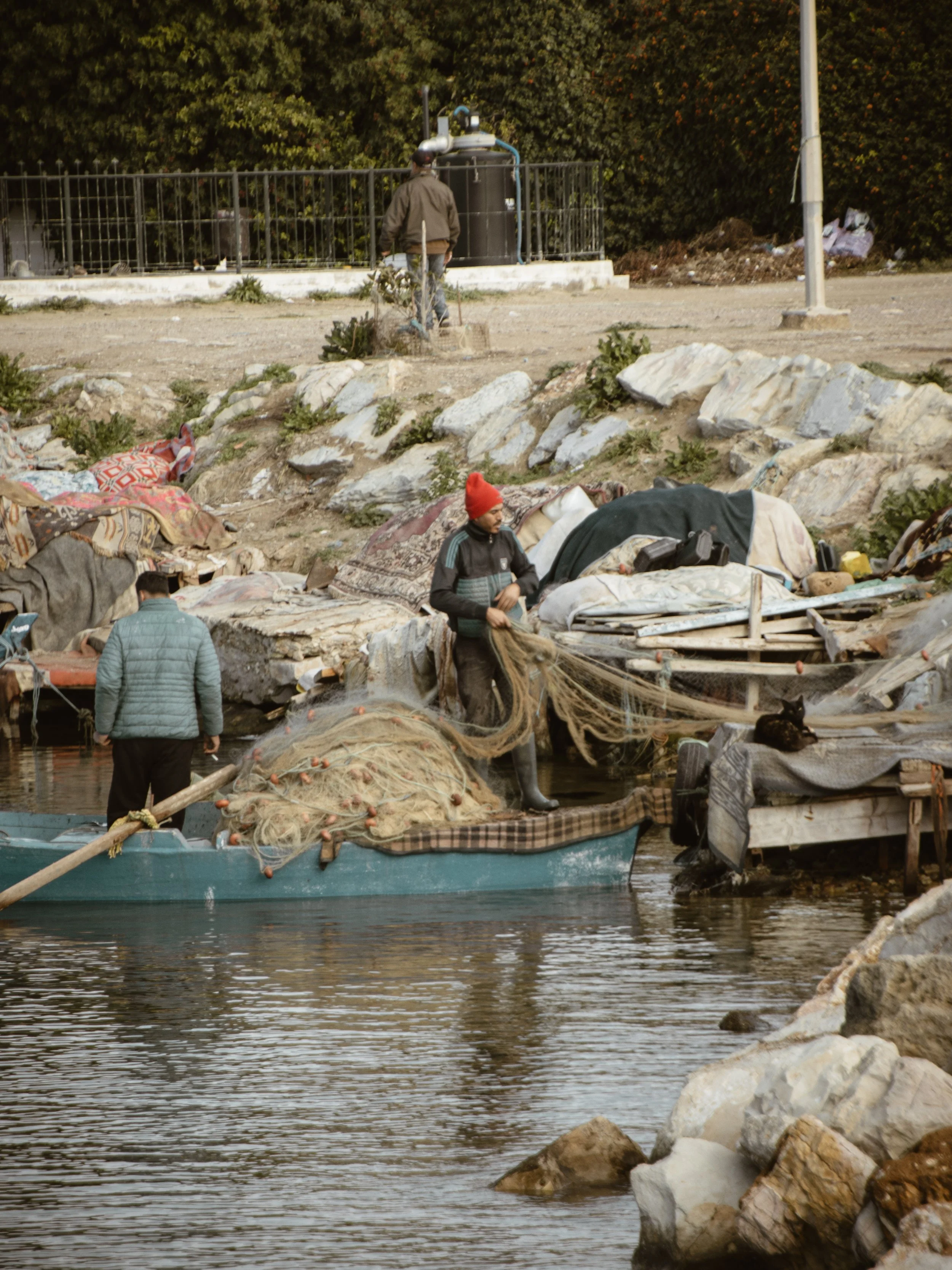 People near rocks and a body of water, using boats with fishing nets, with a background of trees and a fenced structure.
