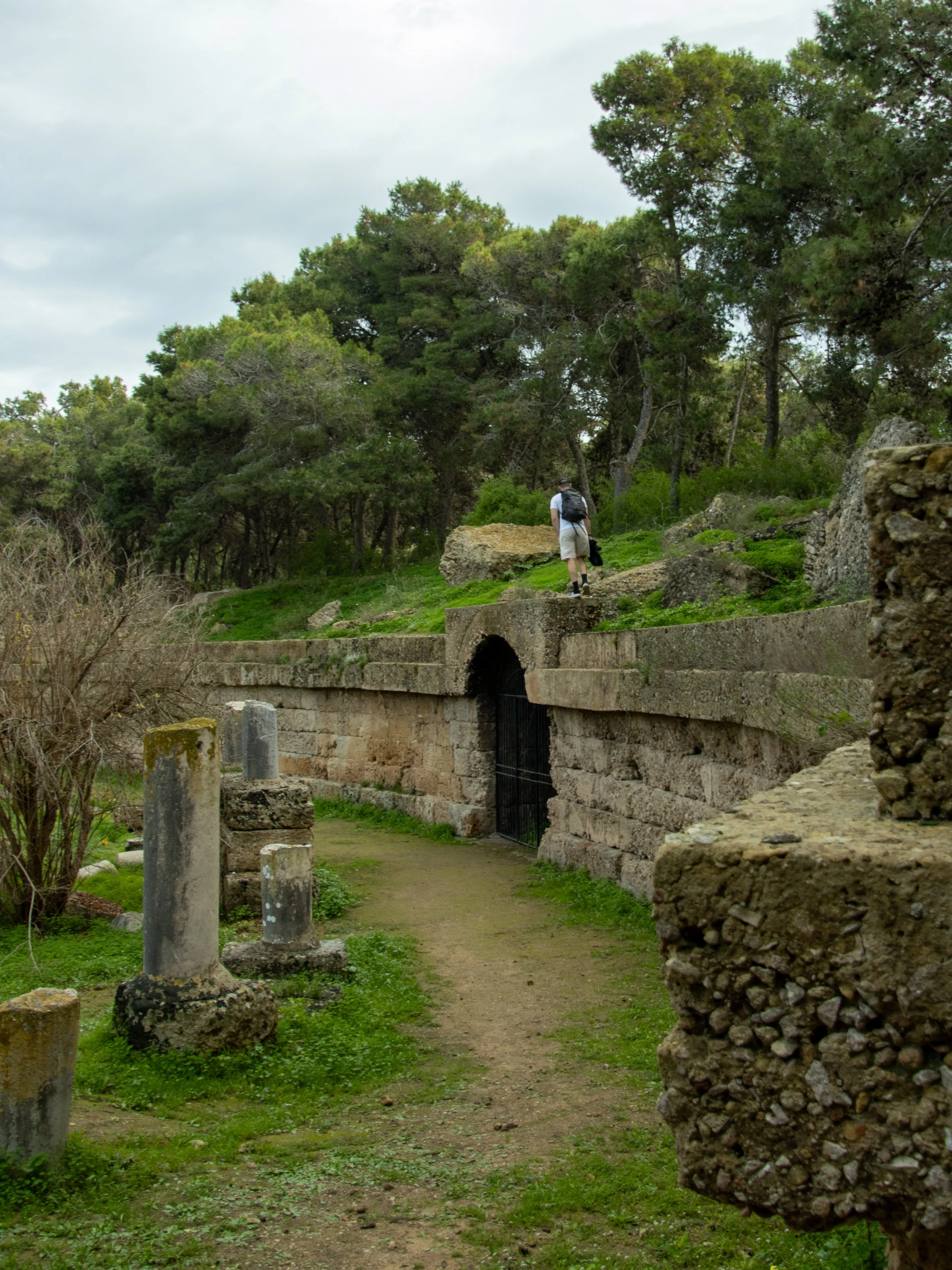 A person hiking on a trail near ancient stone ruins, surrounded by green trees and grass.