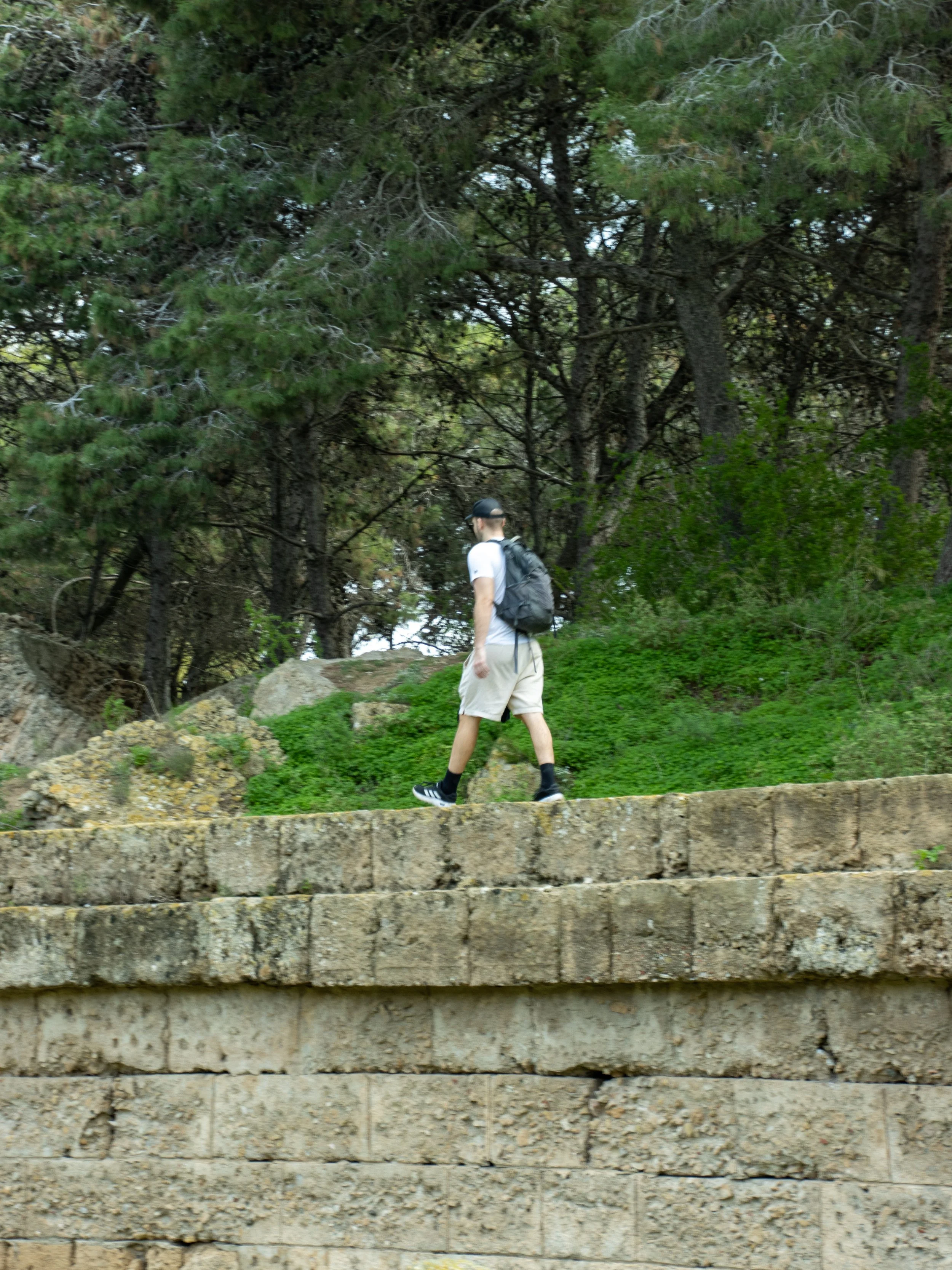 A young man with a backpack walking along a pathway surrounded by trees and green vegetation.