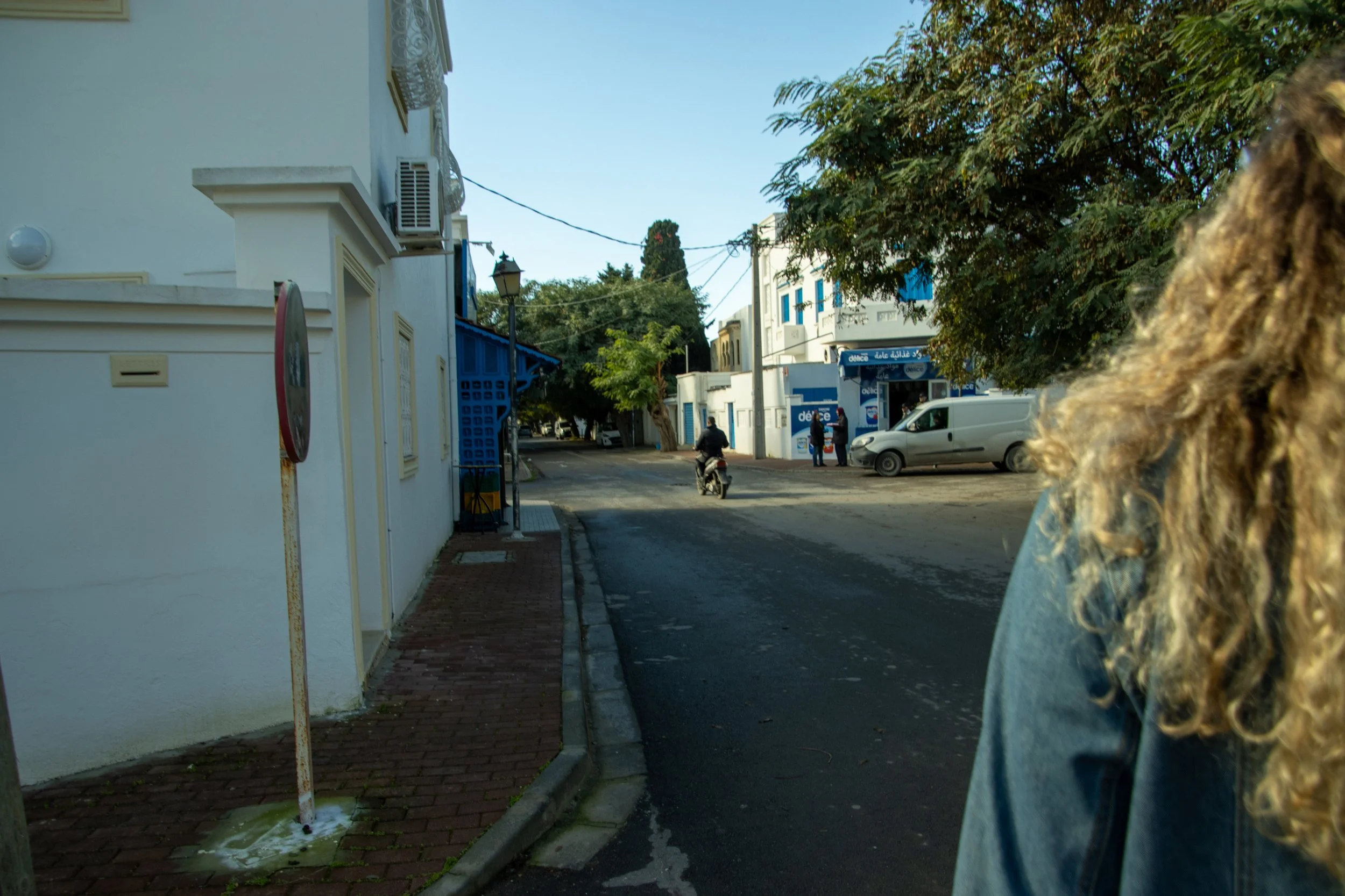 Street scene with white buildings, a person riding a motorcycle, and a few pedestrians. Tree with green leaves and parked cars are visible. The picture is taken from behind a person with curly hair.