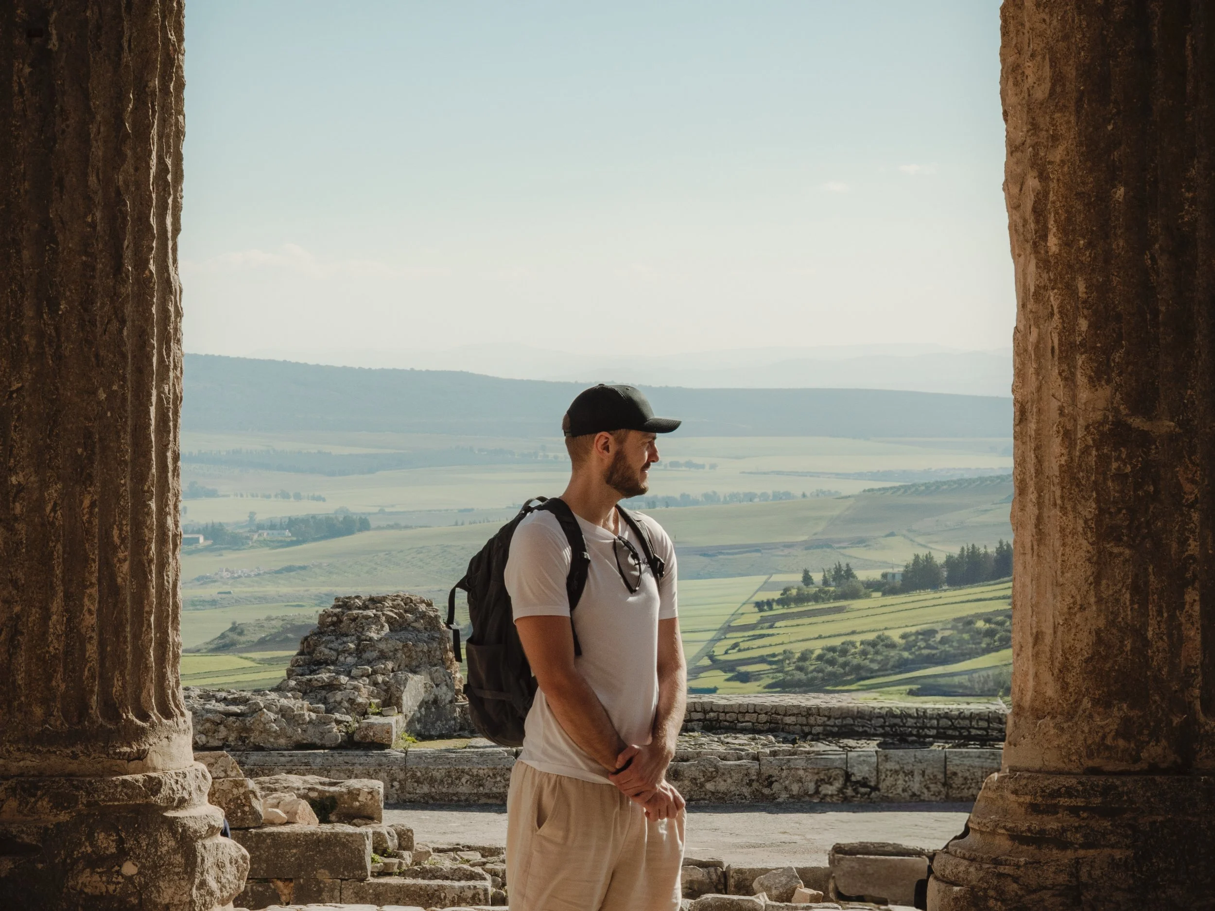 A man with a backpack standing between ancient stone ruins, overlooking a landscape of green fields and distant hills on a clear day.