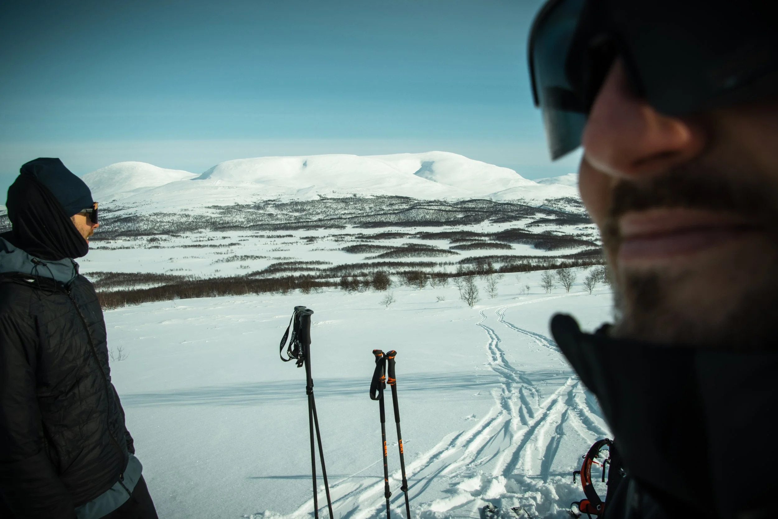 Two people in winter gear standing with ski poles in a snowy landscape, with snow-covered mountains and a clear sky in the background.