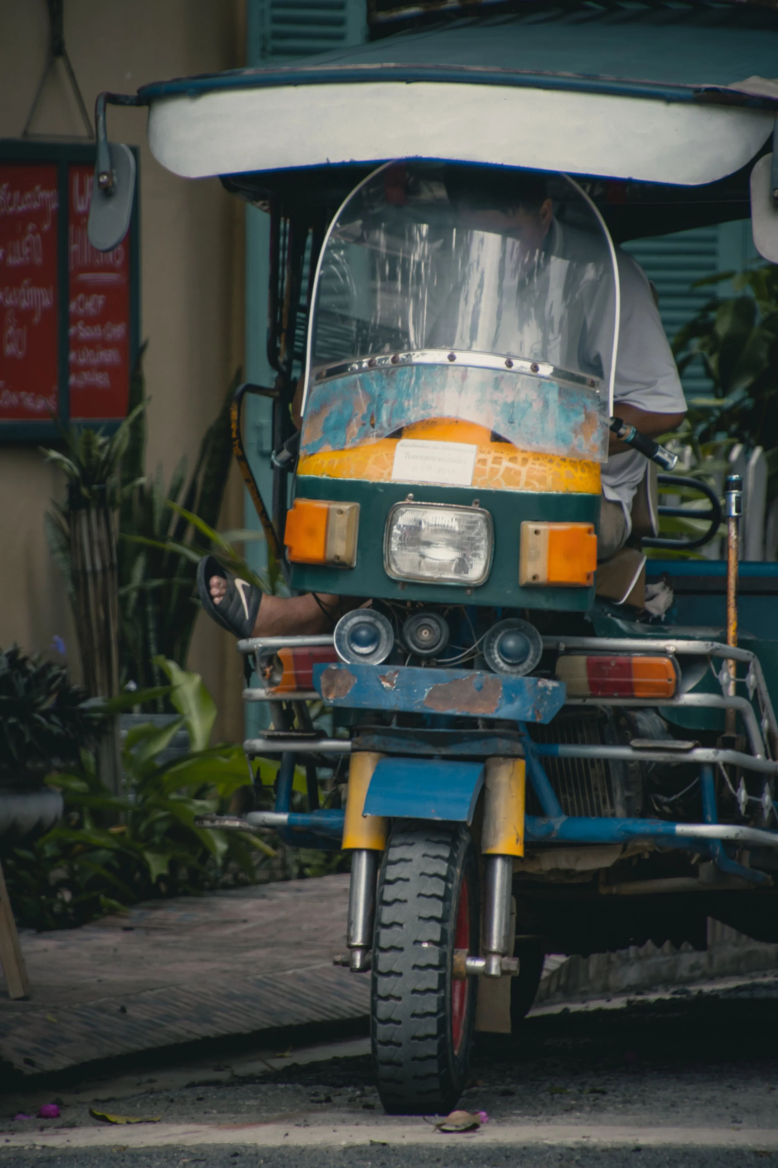 A tuk-tuk rickshaw on a street with a driver partially visible. The vehicle has a blue and yellow color with a transparent windshield, and is parked on a pavement with plants nearby.