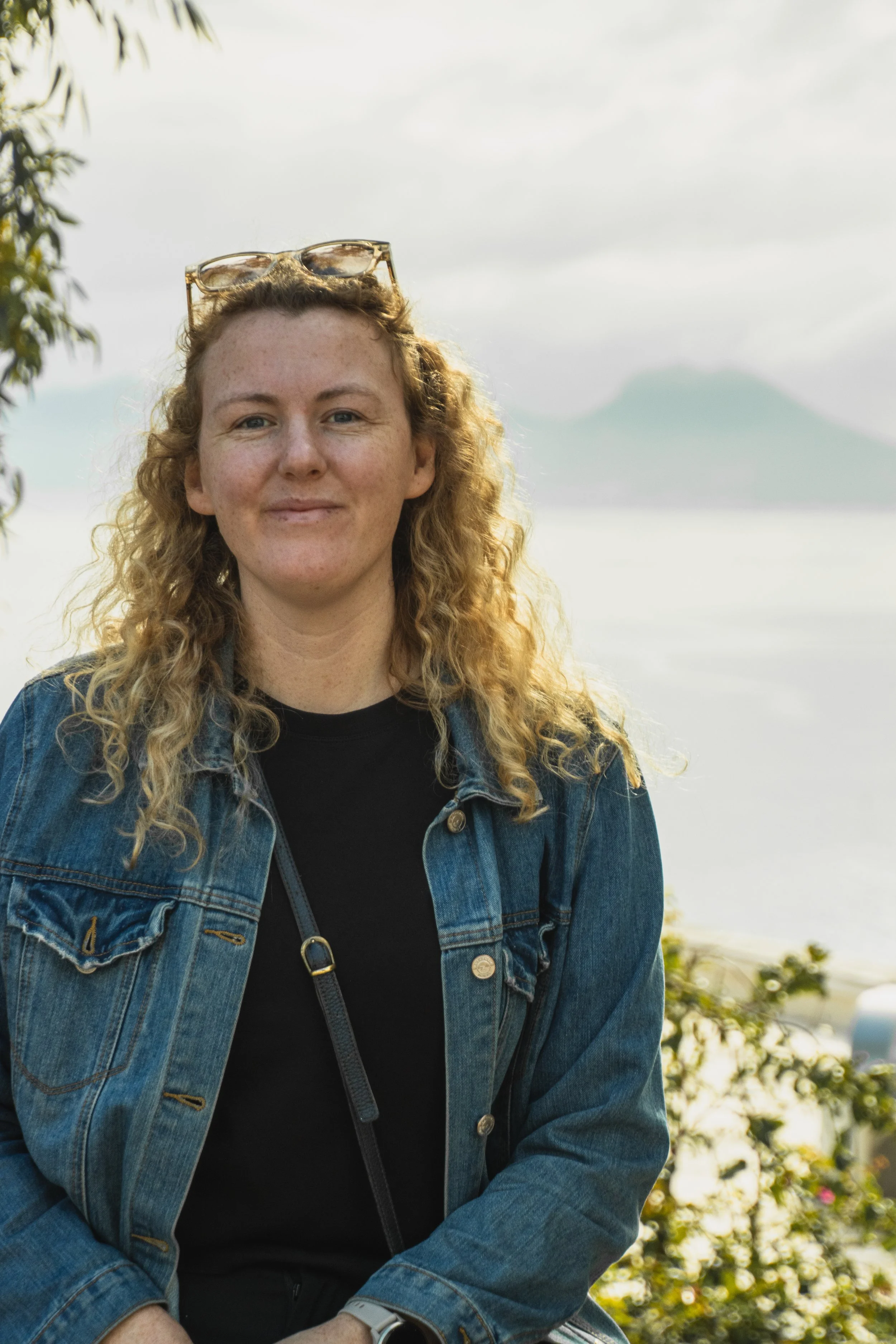 A woman with curly blonde hair and sunglasses on her head, wearing a black shirt and denim jacket, sitting outdoors with a blurred mountain and cloudy sky in the background.