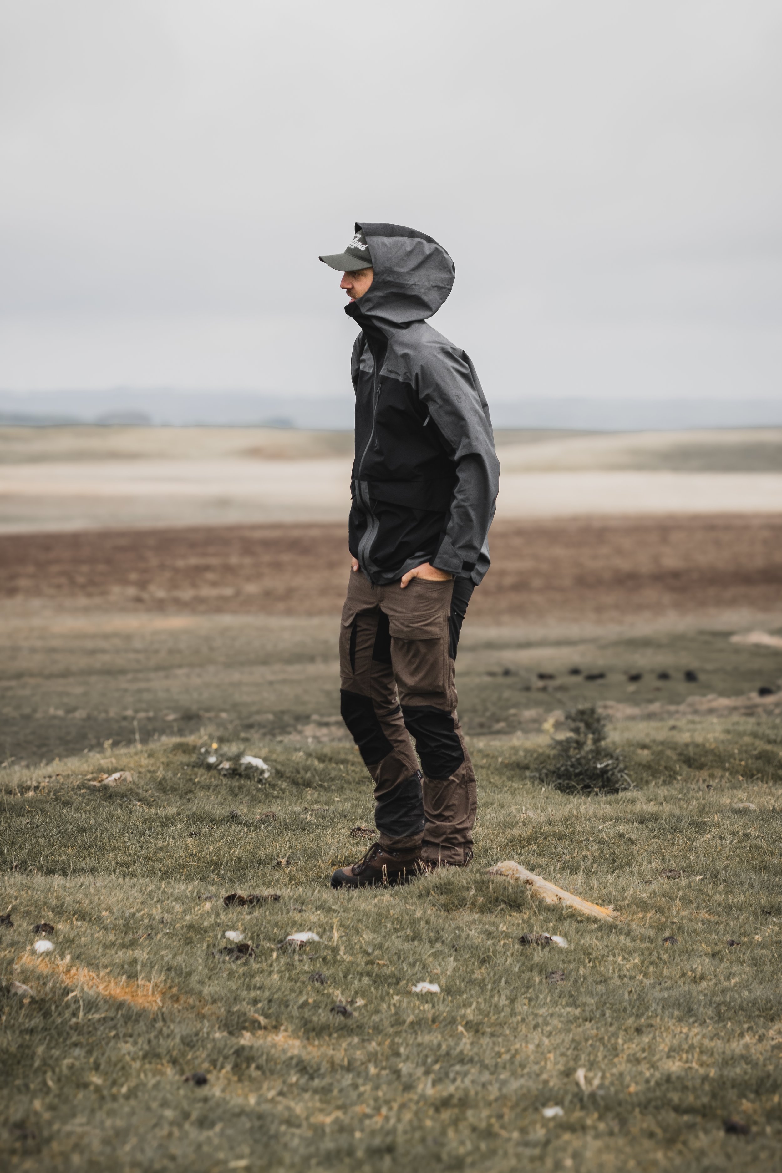 A person wearing a dark rain jacket with the hood up, standing in a grassy area with a cloudy sky and open plain in the background.