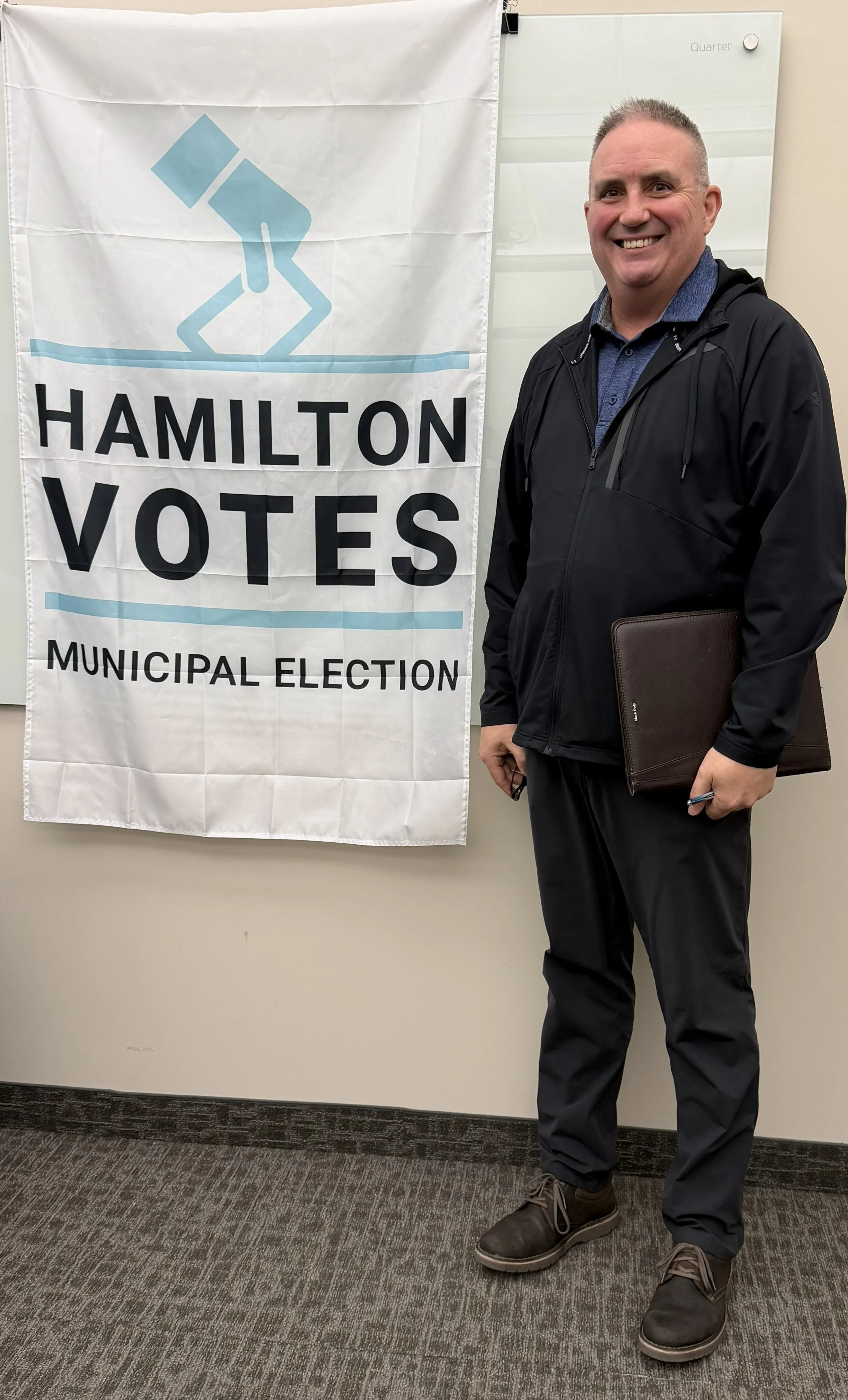 A man standing next to a white banner with the text 'HAMILTON VOTES' and 'MUNICIPAL ELECTION,' holding a folder or notebook, smiling, dressed in a dark jacket and pants, in an indoor setting.
