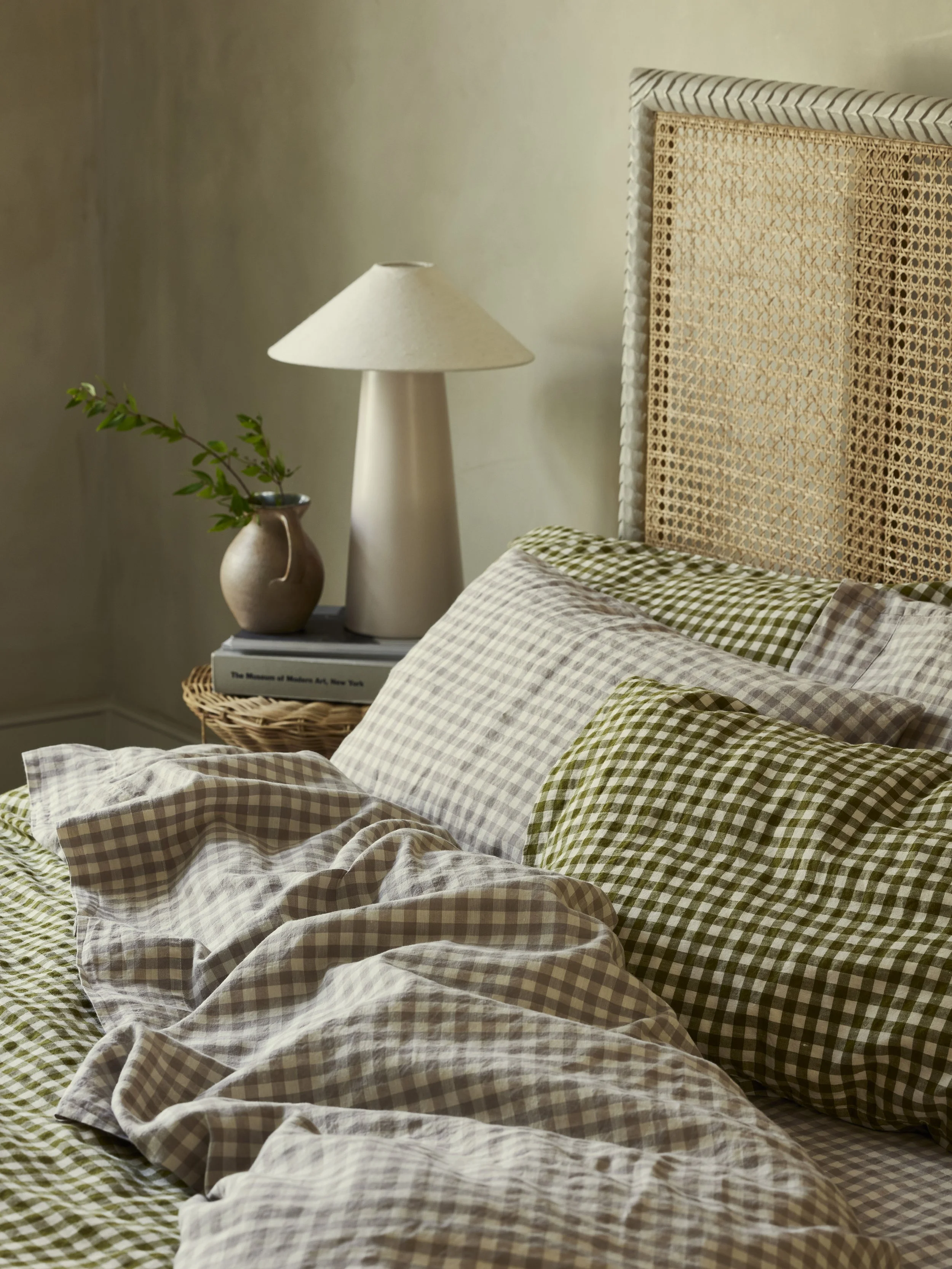 A cozy bedroom scene featuring a bed with green and beige checkered bedding, a rattan headboard, a bedside table with a white lamp, a vase with green leaves, and a stack of books.