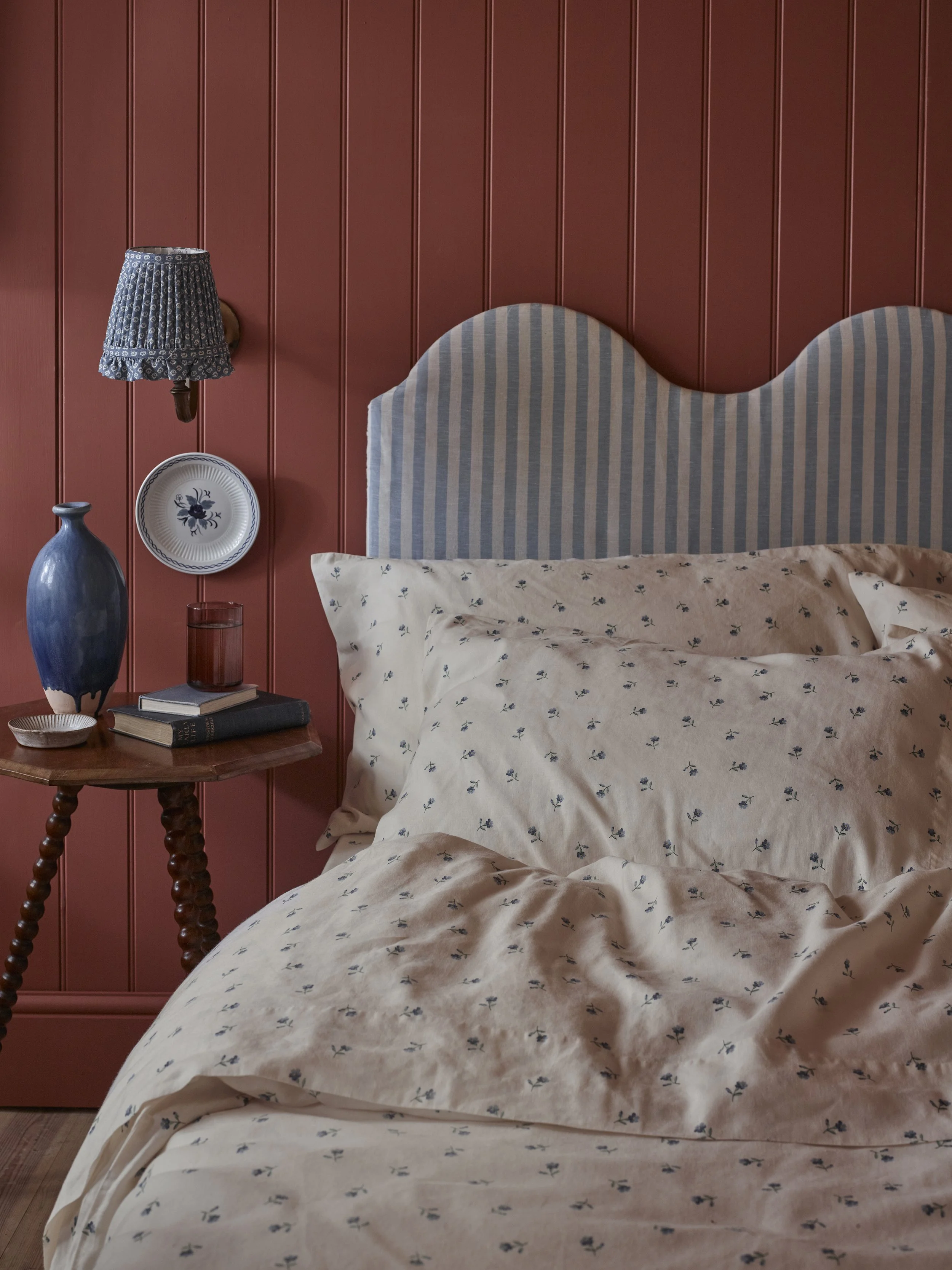 A bedroom with a red wood-paneled wall, a bed with a light-colored, small floral patterned duvet, pillows, a wooden side table with books, a tall blue vase, a small glass, and a vintage wall-mounted lamp with a patterned fabric lampshade.