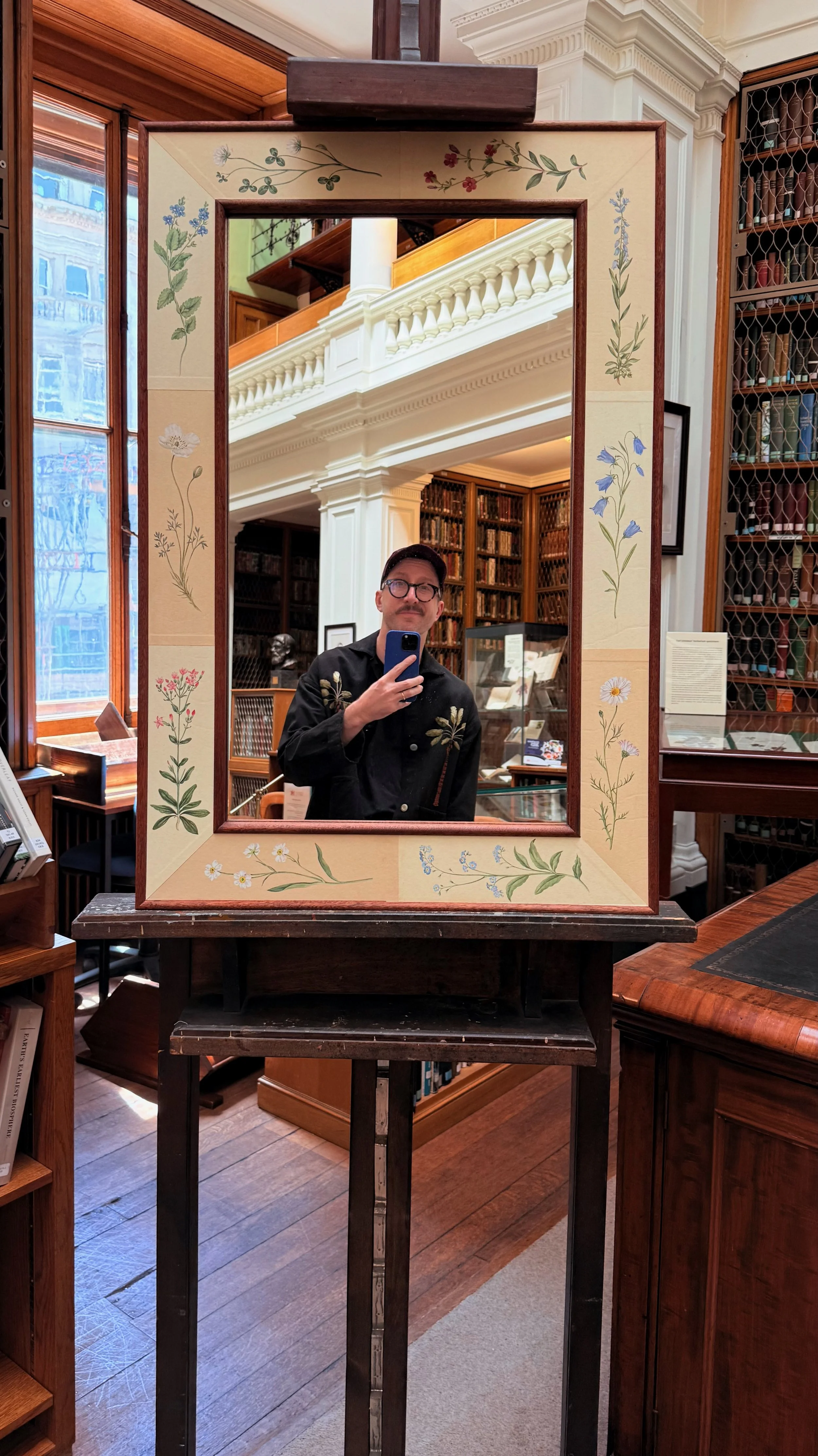 A man taking a selfie in a mirror inside a library or bookstore. The mirror has a floral decorated border, and the surroundings include bookshelves and wooden furniture.