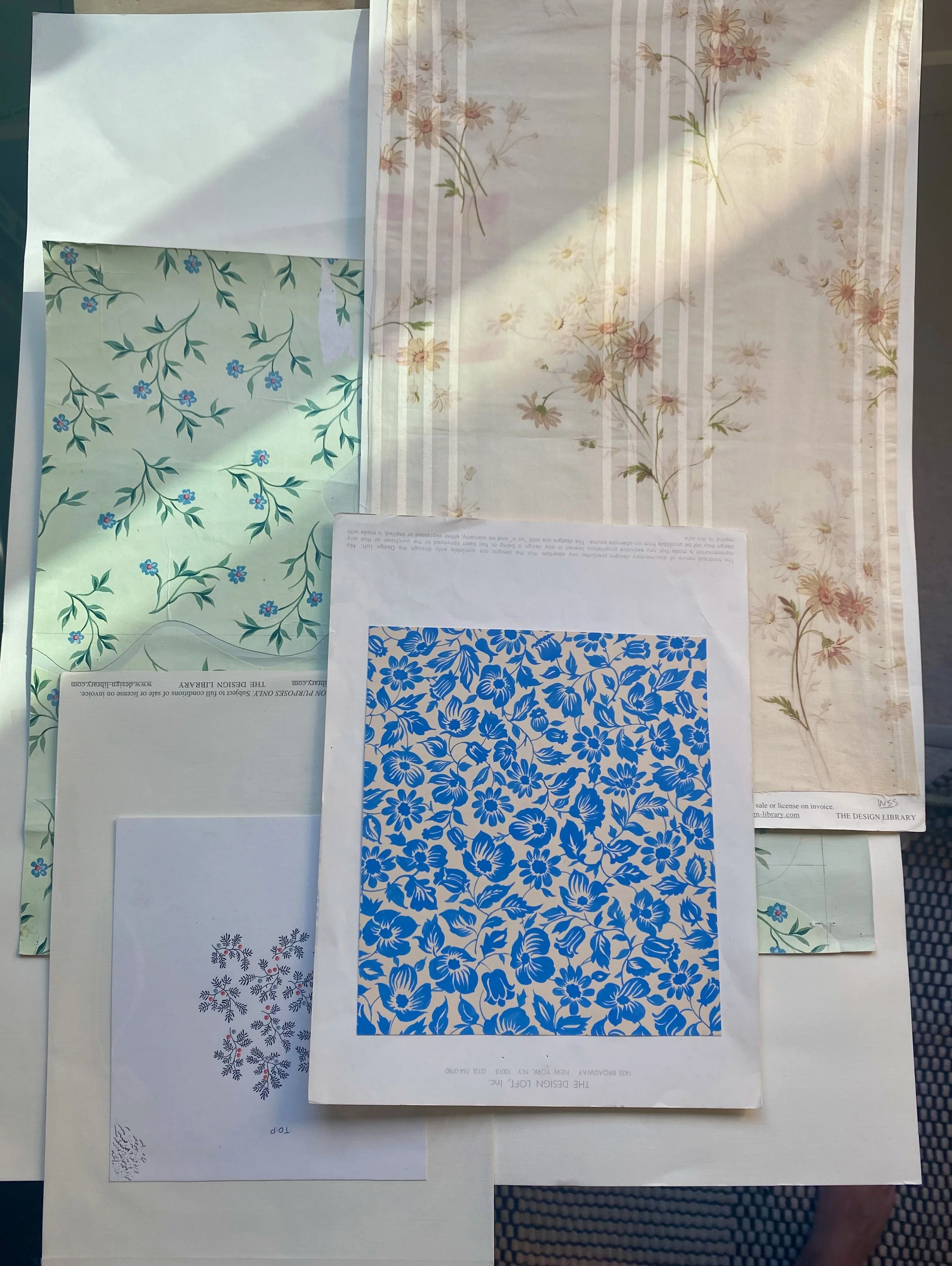 Various patterned wallpapers with floral designs in different colors and styles, displayed on sheets of paper laid out on a surface.