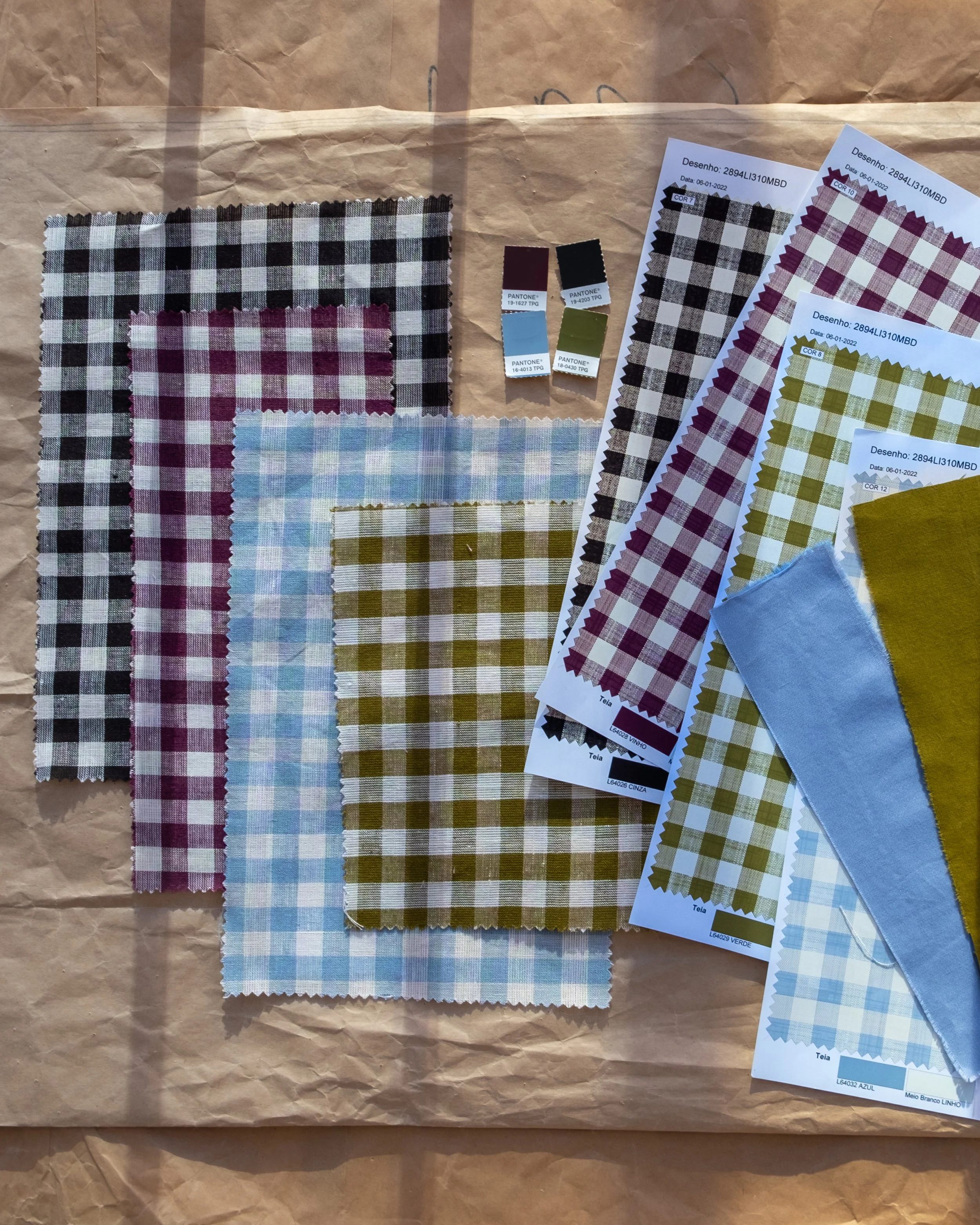 Various fabric swatches with checked patterns in different colors, including black and white, red and white, green and white, and blue and white. Some swatches are attached to printed sheets with fabric code labels and date information. The items are laid out on a crumpled brown paper background.