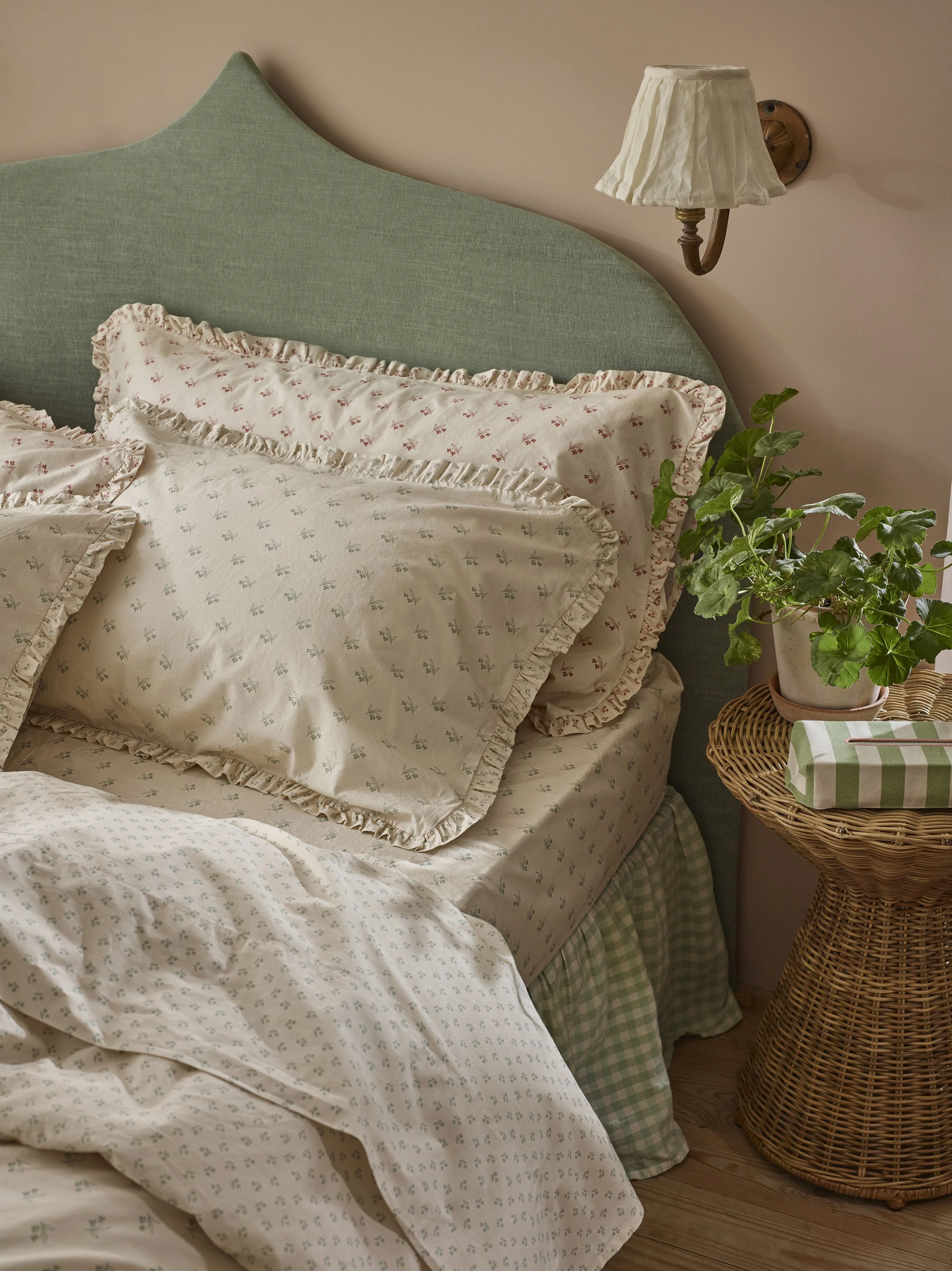 A neatly made bed with floral ruffle-edged pillowcases, a green headboard, a bedside table with a potted plant, and a wicker side table with a box and a wooden floor.