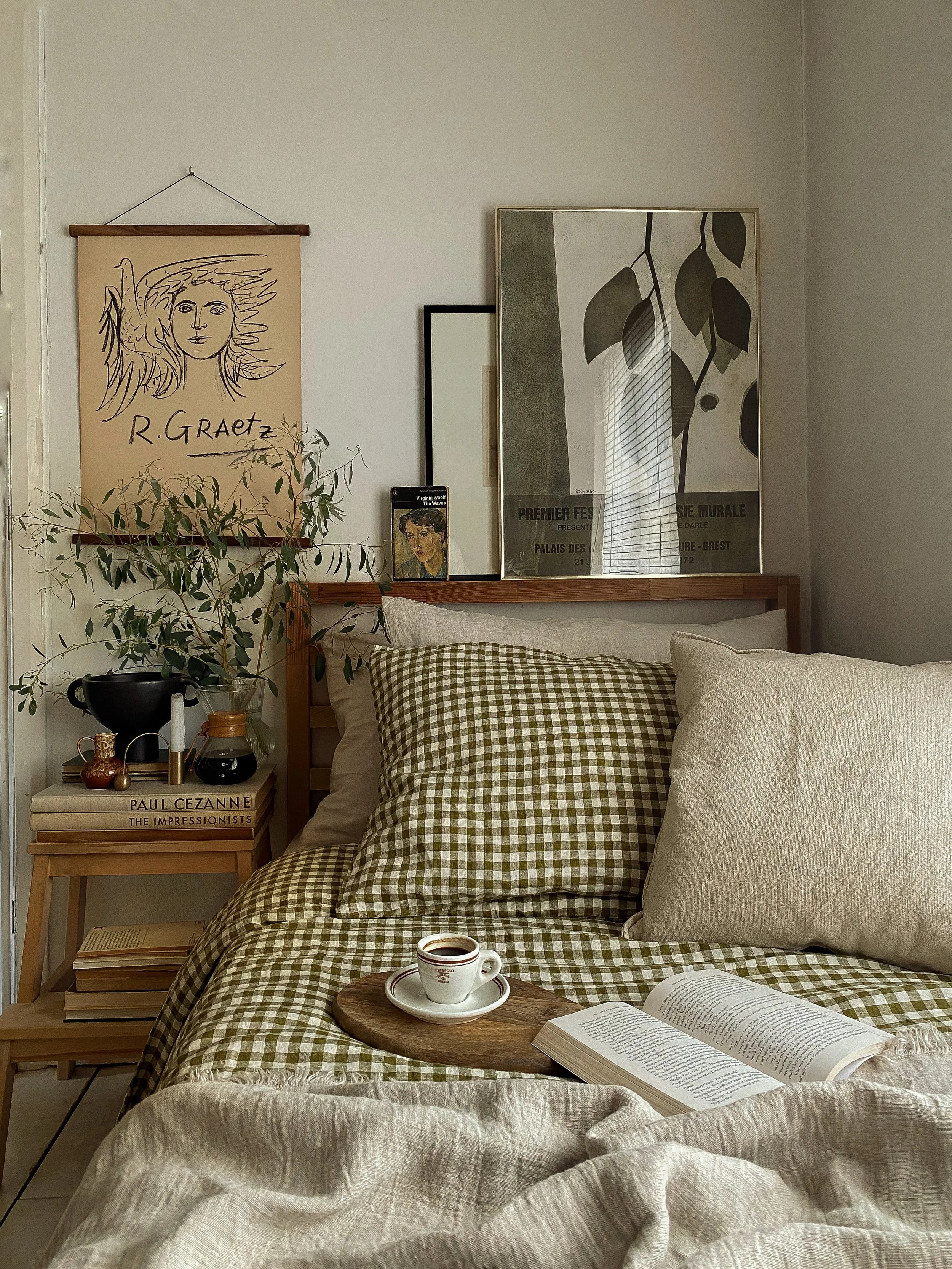 A cozy bedroom with a bed covered in a checkered bedspread and a large beige pillow. A wooden tray with a cup of coffee and an open book is on the bed. There is a nightstand with books, a plant, and decorative items. Framed artwork, including a line drawing of a woman and a leaf print, hang on the wall. A mirror and some smaller frames are also visible.