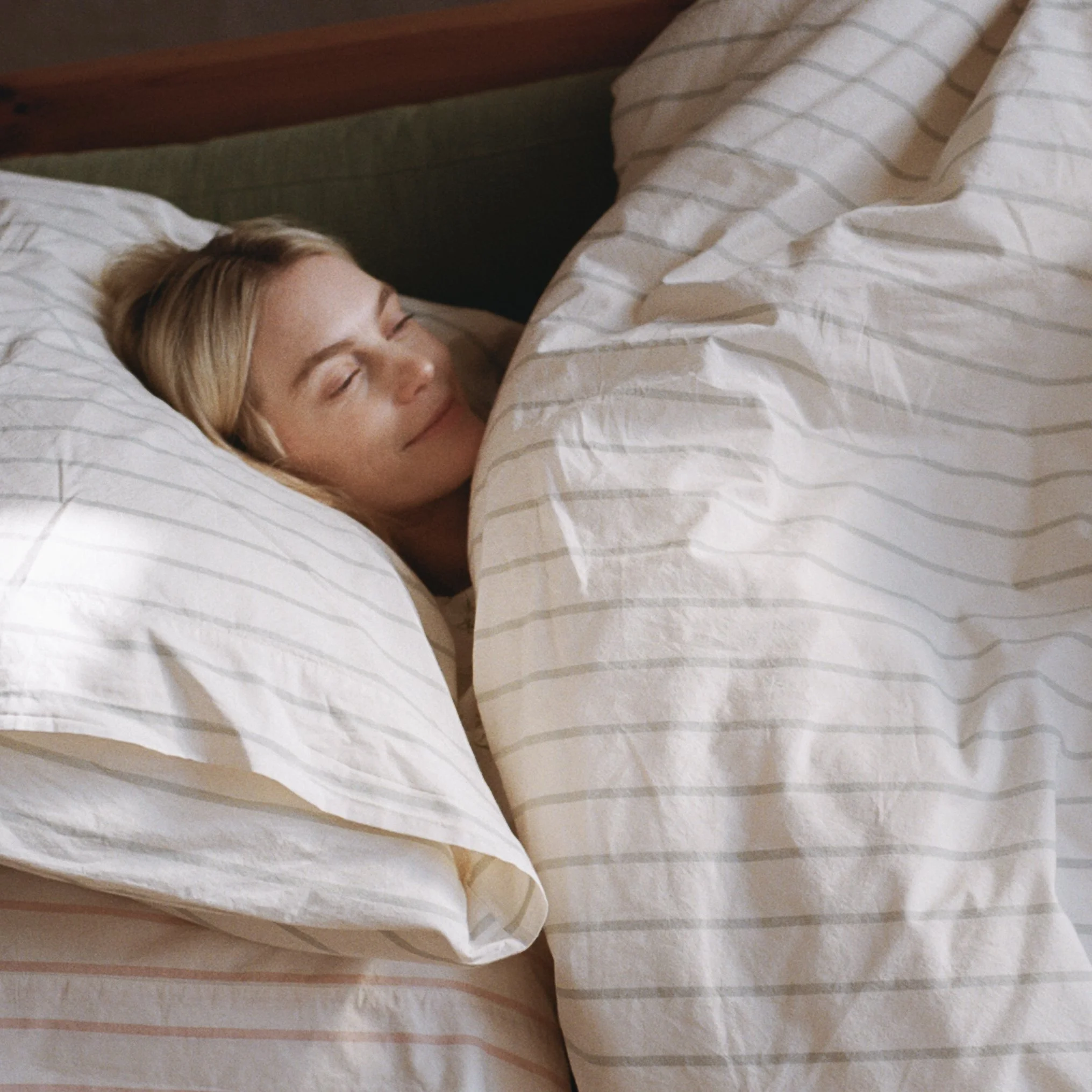 A woman with blonde hair sleeping peacefully in bed, covered with a cream-colored striped duvet and lying on a white pillow, in a cozy bedroom setting.