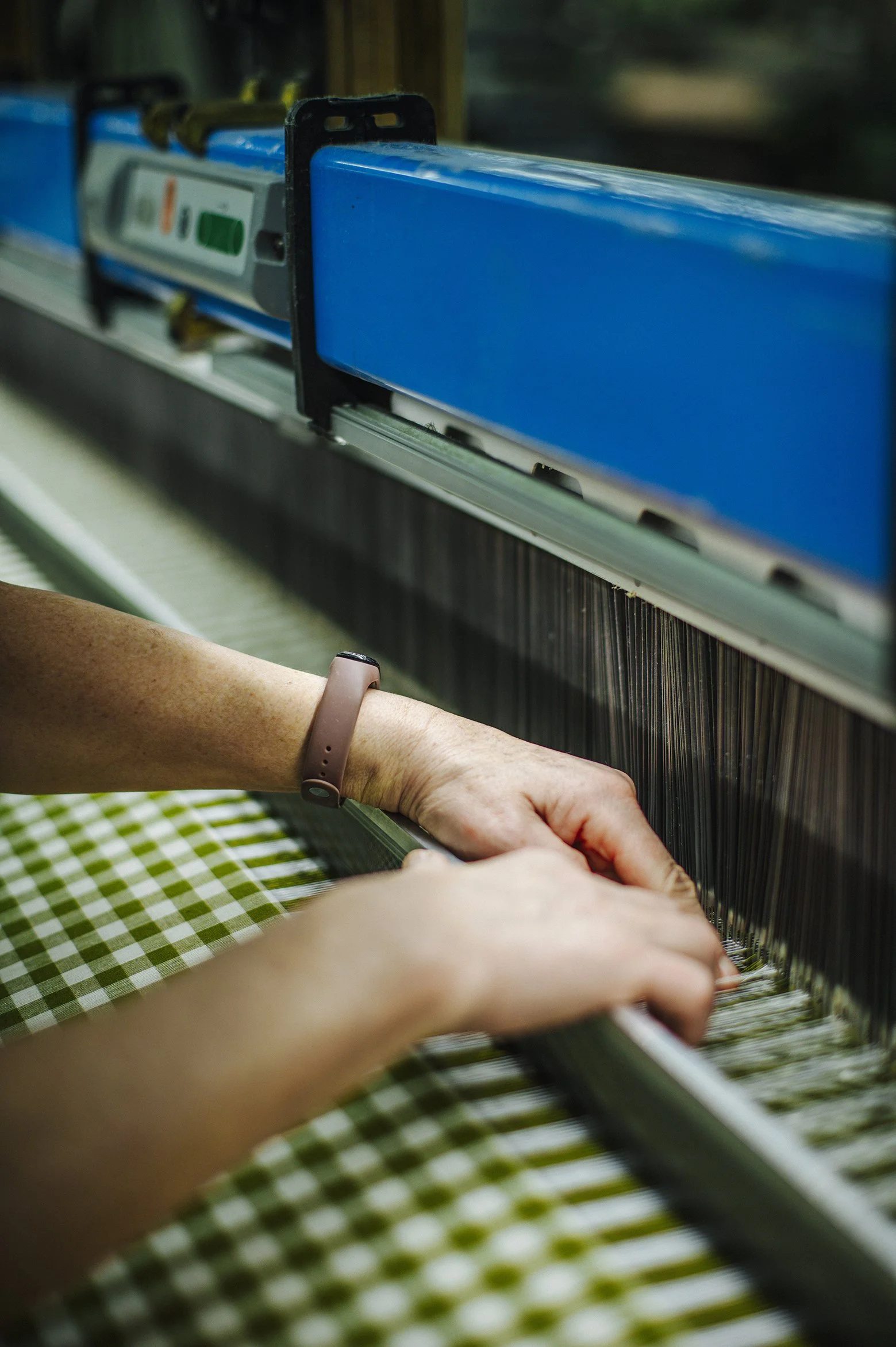Close-up of a person's hands working on a textile weaving machine, with green and white checked fabric in the foreground.