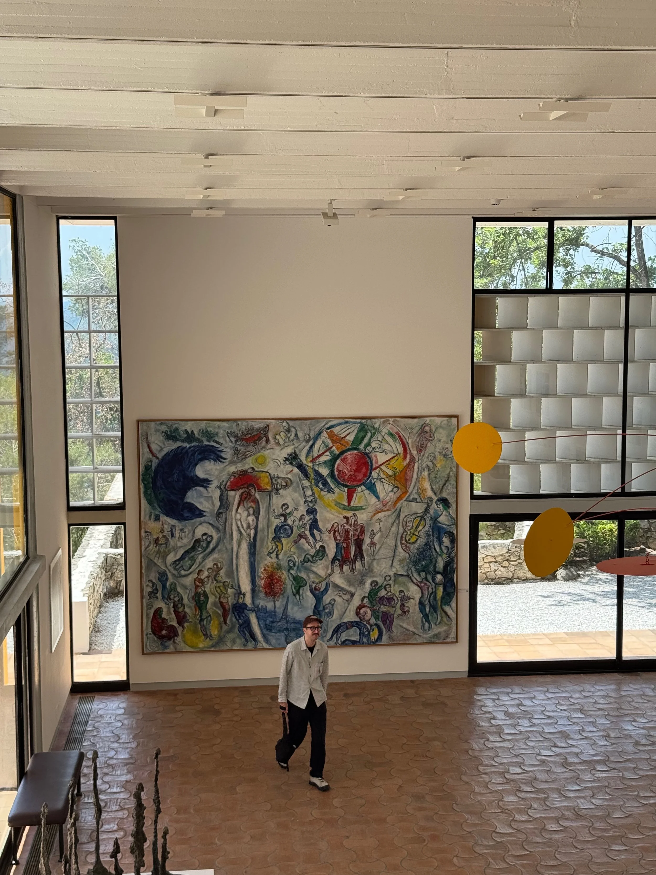 Interior view of an art gallery with large abstract painting on white wall, windows showing trees outside, and a man standing near the center of the room.
