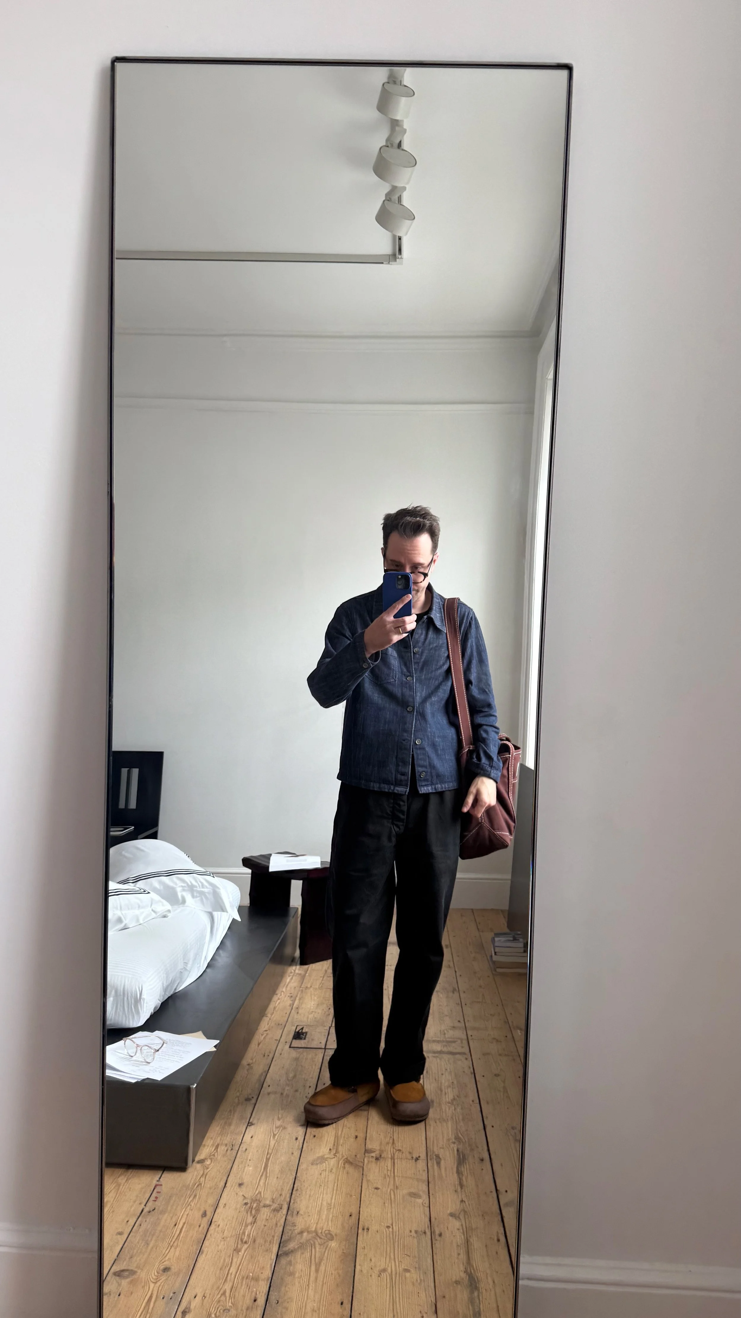 A man taking a mirror selfie in a bedroom, wearing a dark jacket, black pants, yellow shoes, glasses, and carrying a brown shoulders bag. The room has a bed with white sheets, a bedside table with papers and glasses, and wooden flooring.