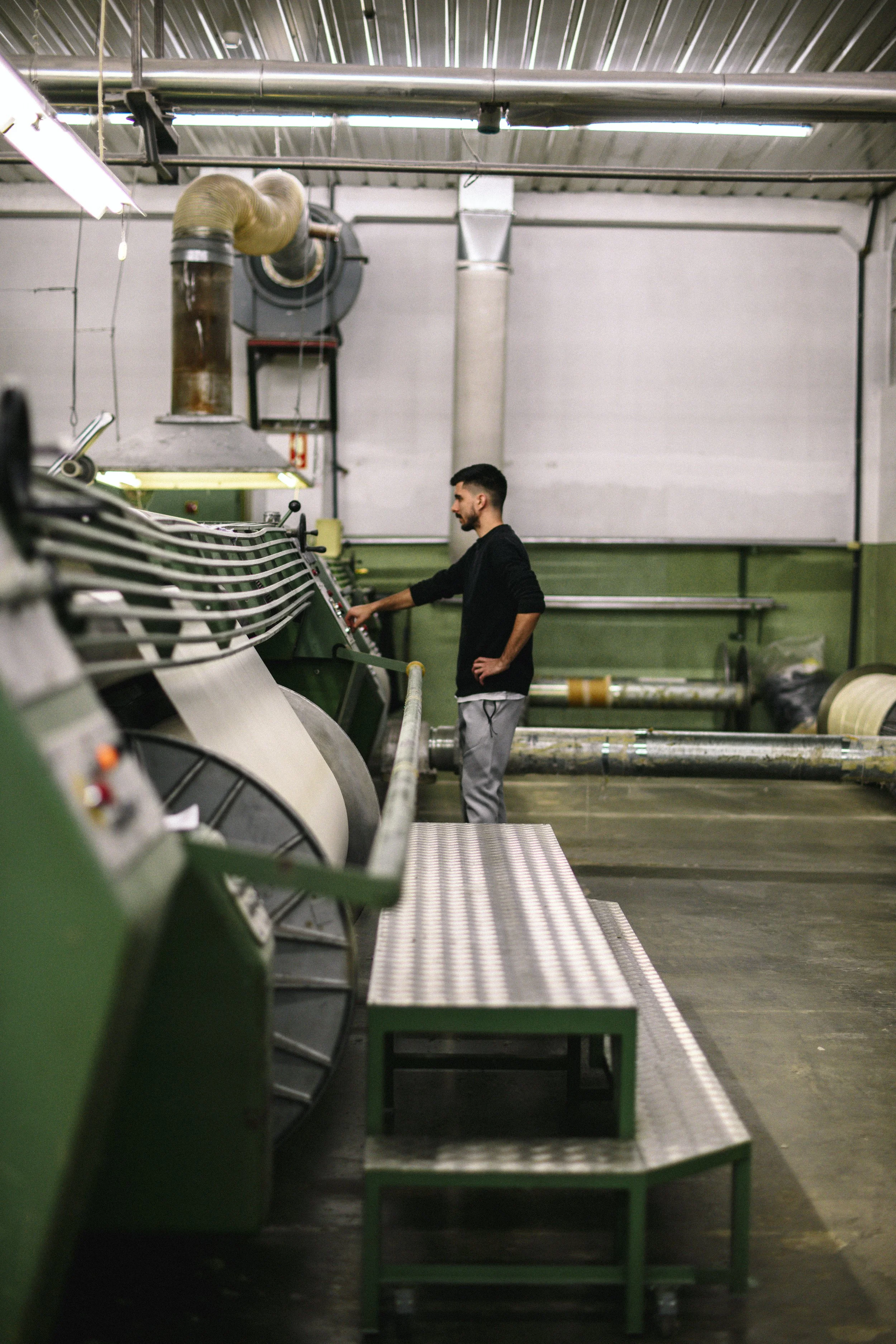 A man wearing a black long-sleeve shirt and gray pants operating a machine in an industrial factory setting with green and metal machinery, pipes, and ventilation ducts overhead.