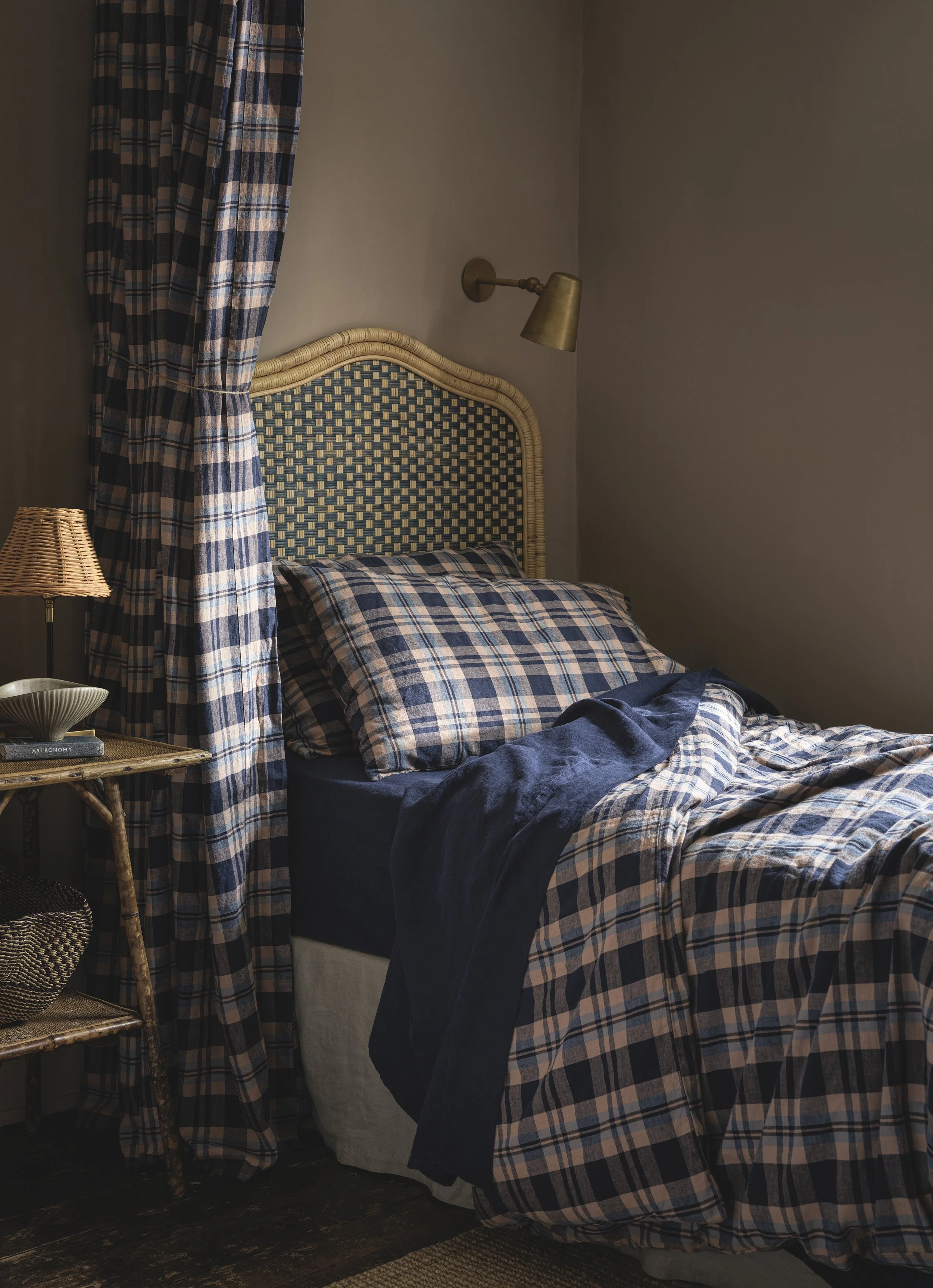 A cozy bedroom with a bed that has a rattan headboard and plaid bedding in shades of blue and beige. There is a plaid curtain matching the bedding on the left side, a small bedside table with a book titled "Astronomy" and a decorative bowl, and a brass wall-mounted reading lamp above the bed. The room has dark wood flooring and neutral-colored walls.