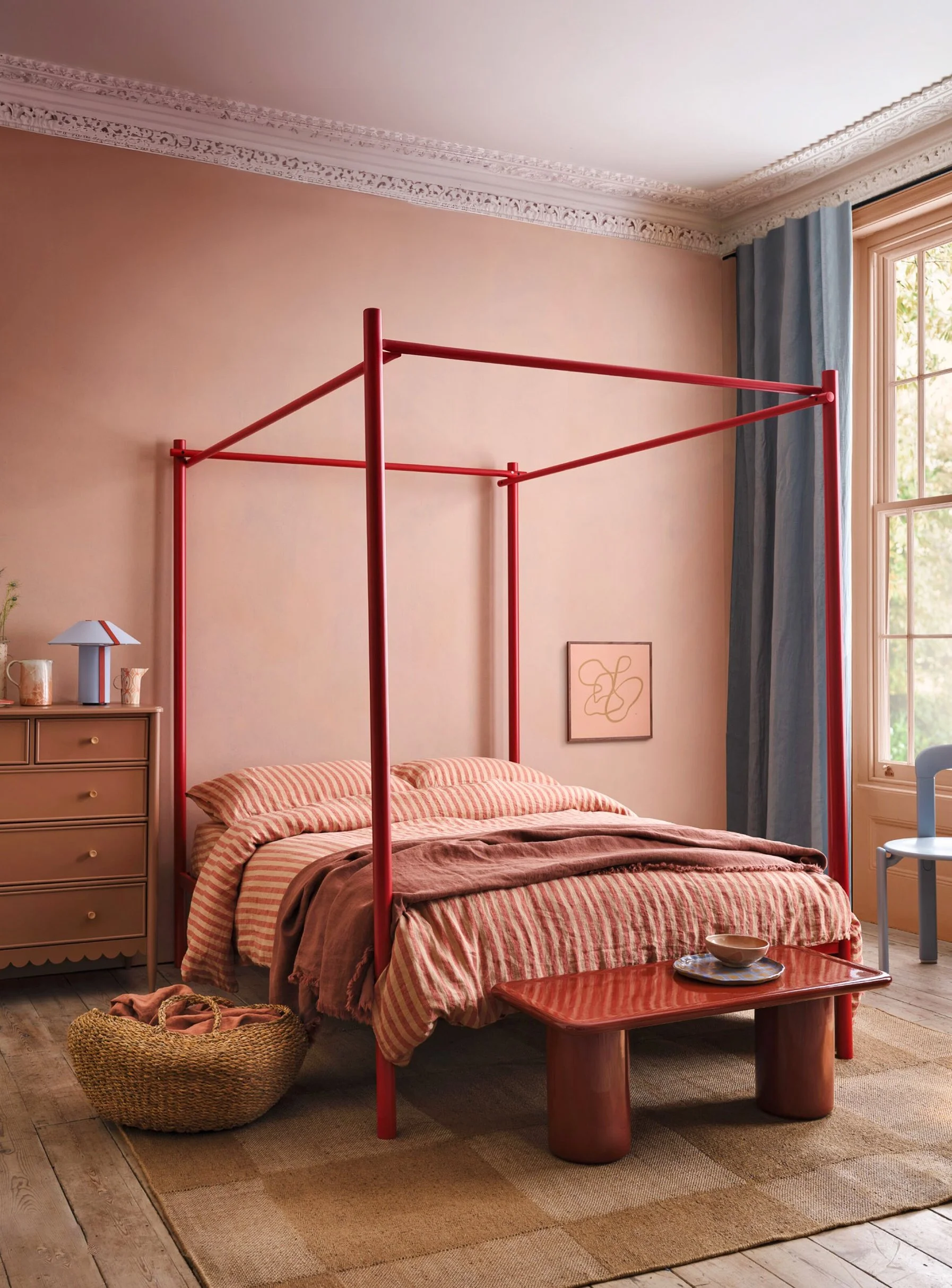 A bedroom with a pink four-poster bed, pink and white striped bedding, a beige dresser with a blue and white lamp, blue curtains, a large window, a framed artwork, a wooden coffee table with a blue plate and a bowl, a woven basket with clothes, and a beige and brown rug on a wooden floor.
