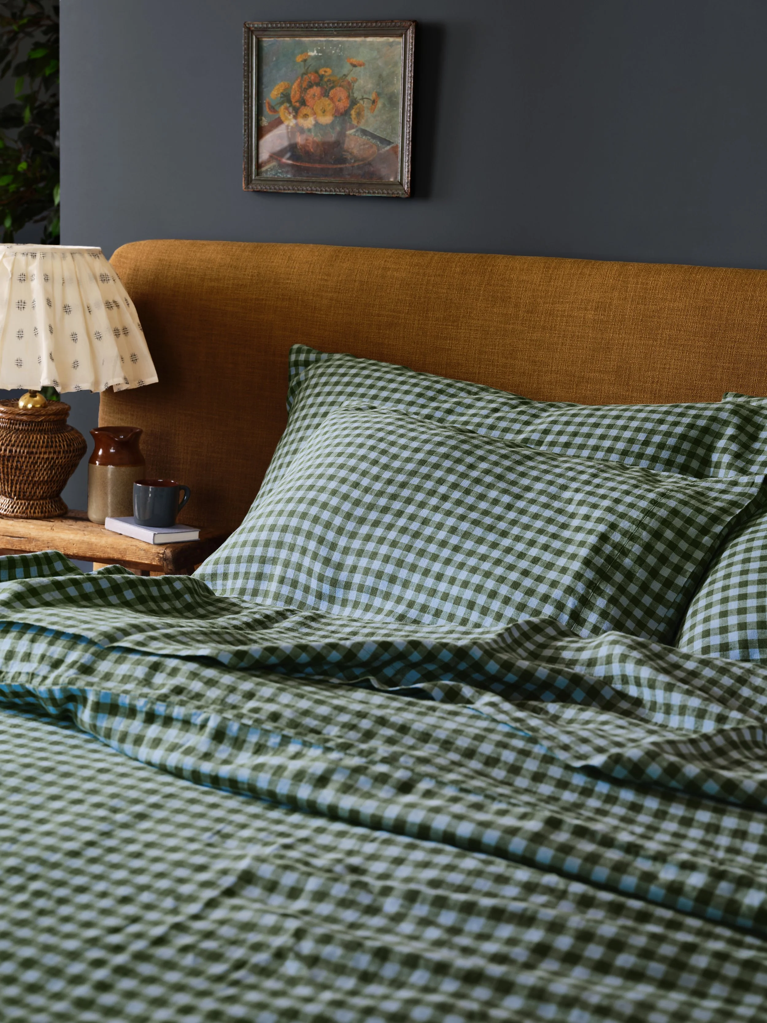 A bedroom scene with a bed covered in green and white checkered bedding, a pillow matching the bedding, a wooden bedside table with a book, a gray mug, and a ceramic vase, a vintage table lamp, and a framed floral painting on the blue wall.