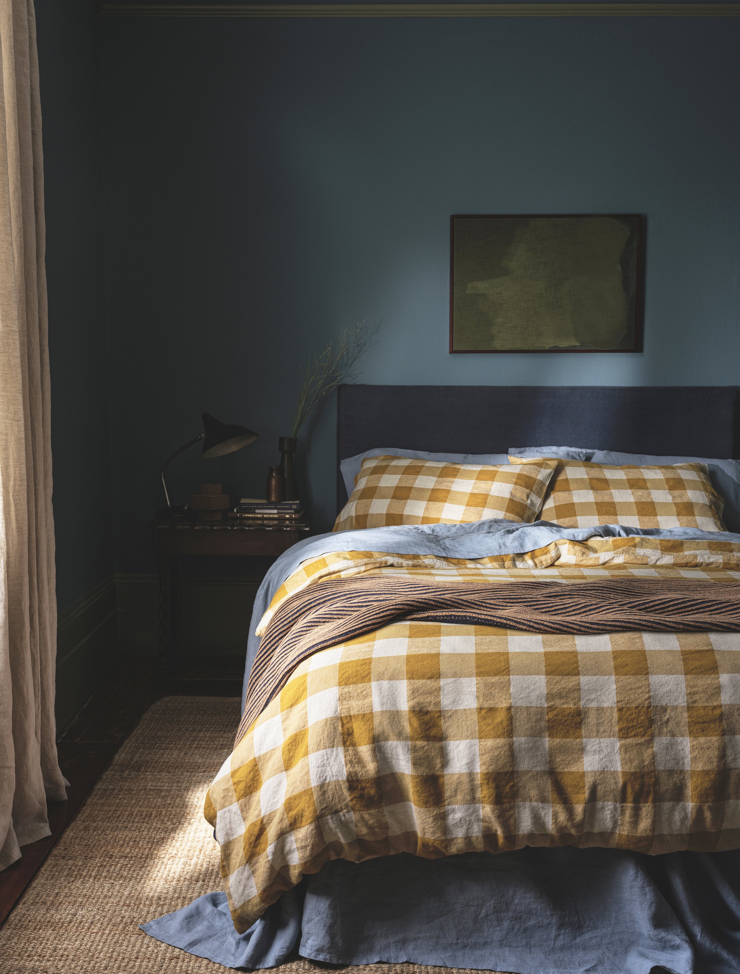 A bedroom with a bed that has a blue headboard, yellow checkered bedding, and a blue bed skirt. To the left, there is a nightstand with a black desk lamp, some books, and a decorative item. There is a large, framed picture on the blue wall above the bed. A tall vase with branches is on the nightstand. The floor is carpeted, and sunlight is coming in from a window with beige curtains on the left side of the image.